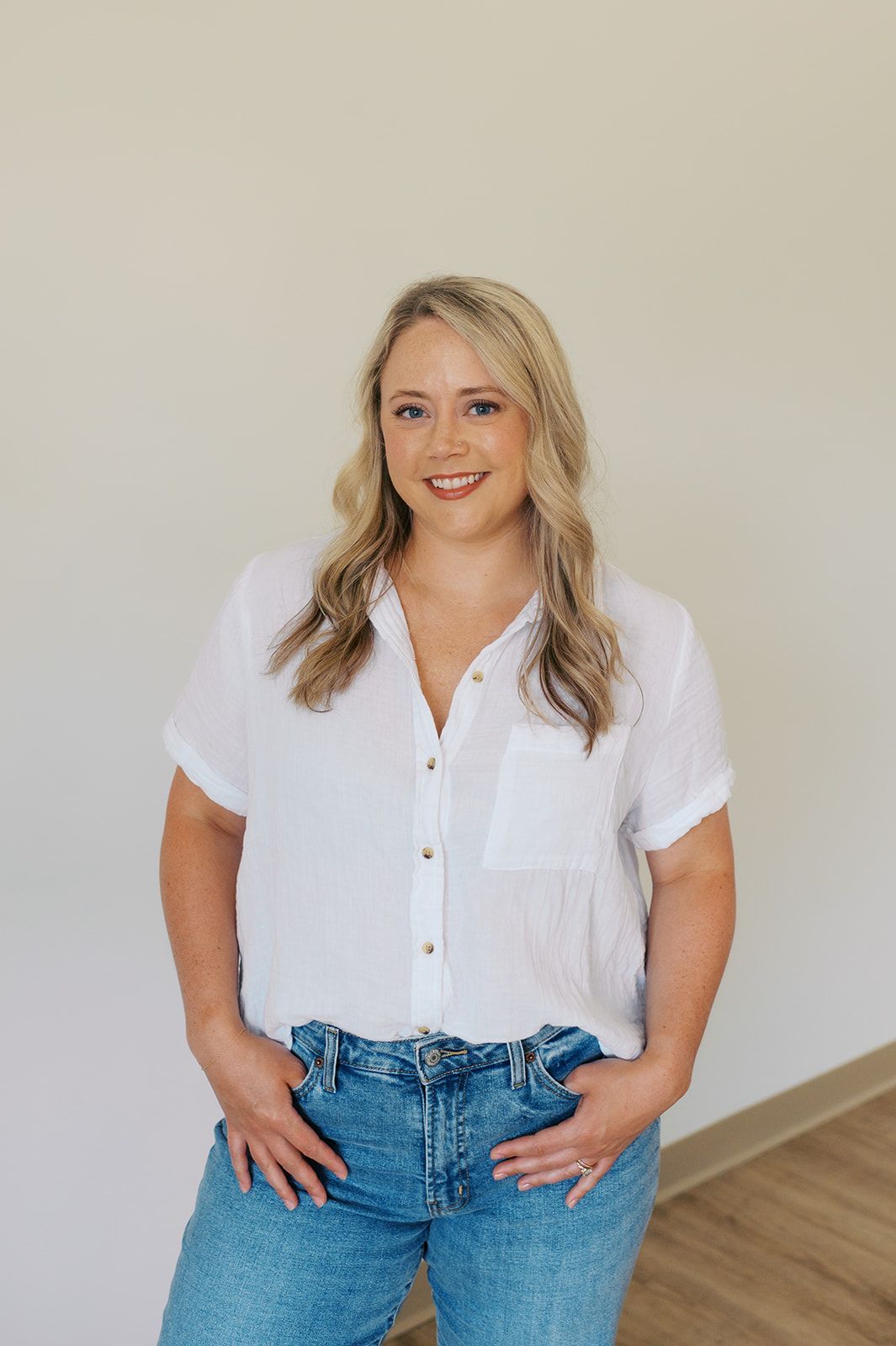 A person with shoulder-length blonde hair, wearing a white button-down shirt and blue jeans, standing against a wall.