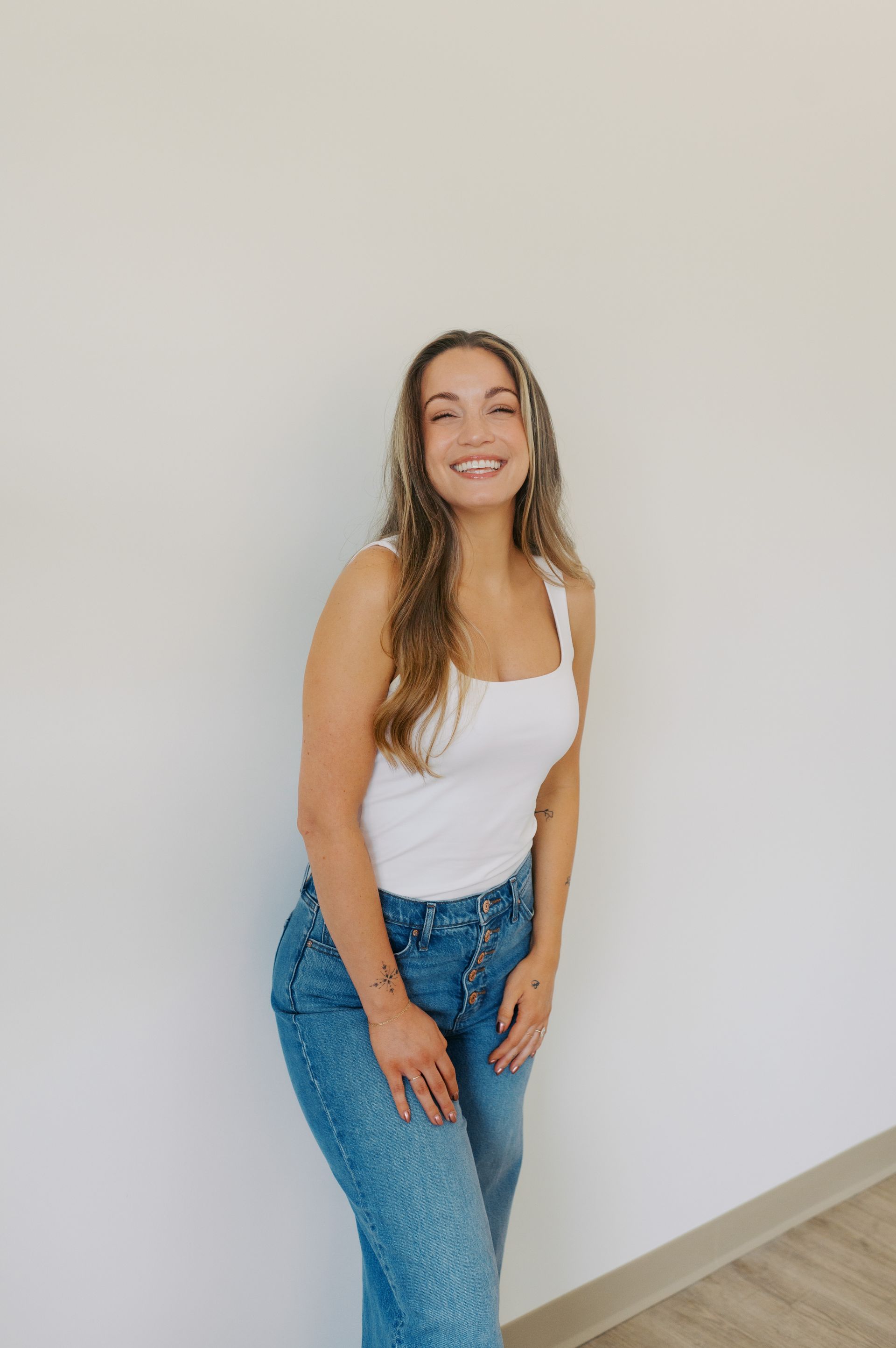 A person with long brown hair, wearing a white tank top and blue jeans, smiles while posing against a plain wall.