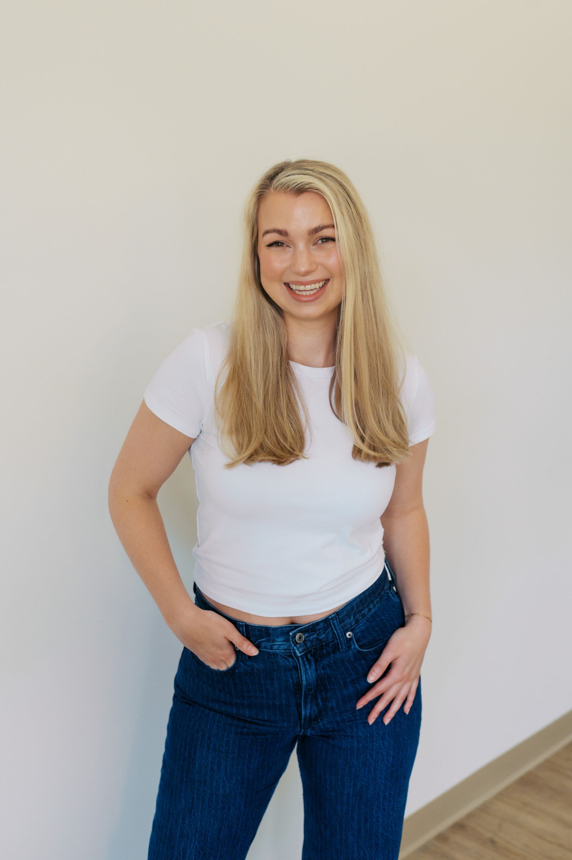 Smiling person in a white tie-strap top and blue jeans standing in front of white shelves filled with products.