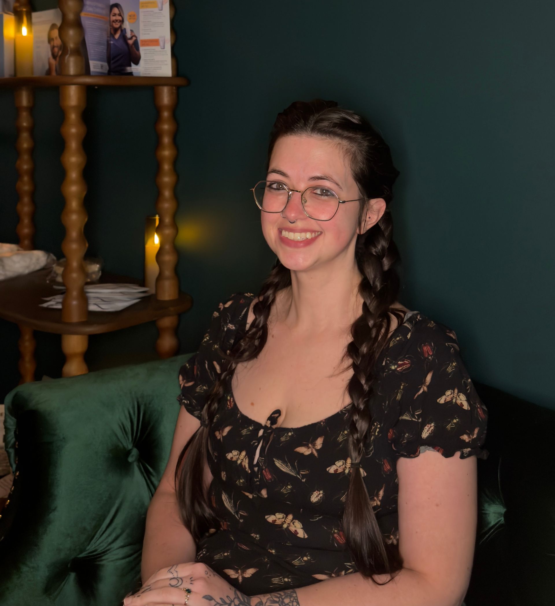 Smiling person with glasses and braided hair wearing a dark patterned dress, sitting in front of a shelf.