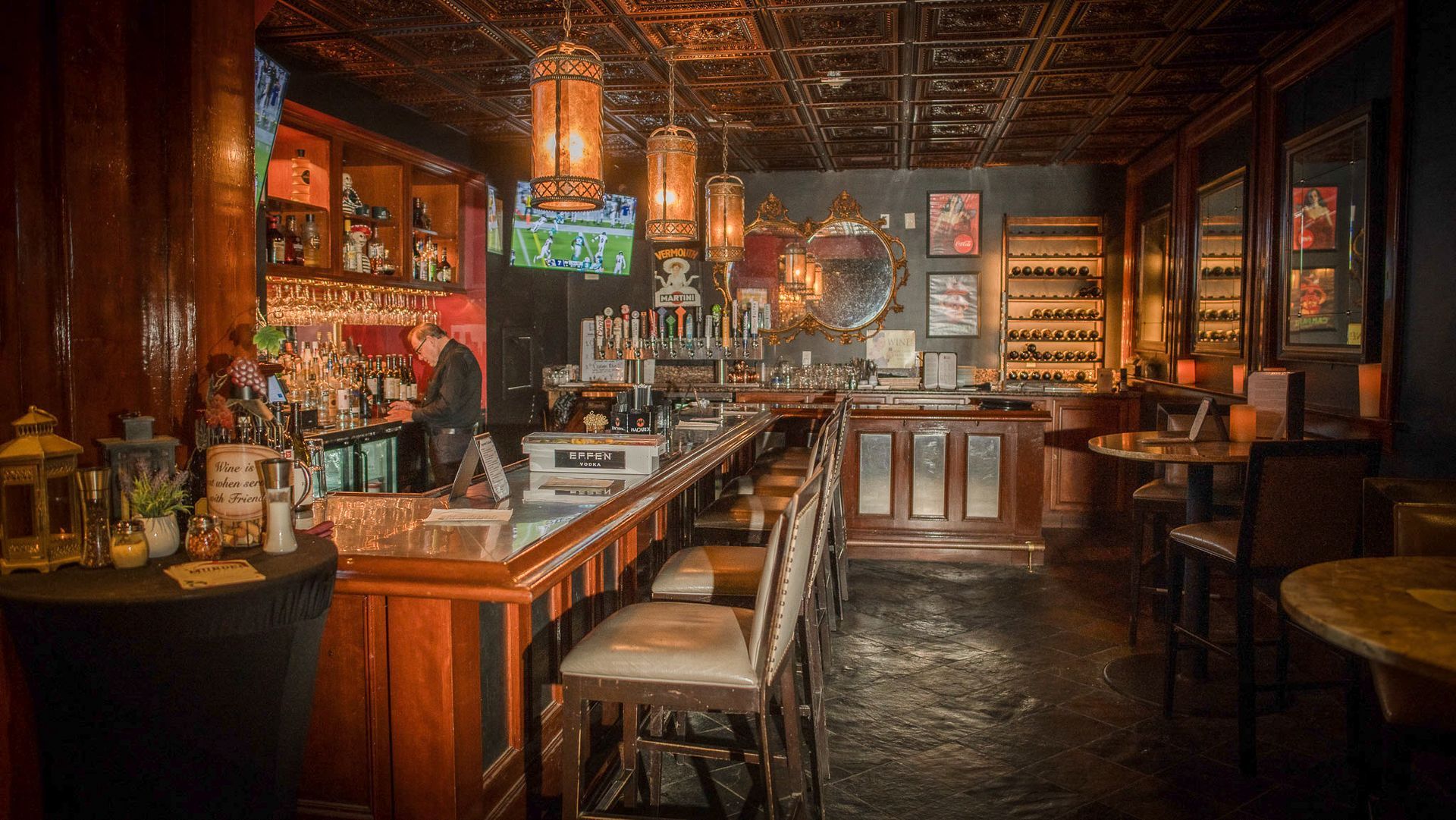 Interior view of a dimly lit bar with a long wooden counter, stools, and ornate ceiling lights.