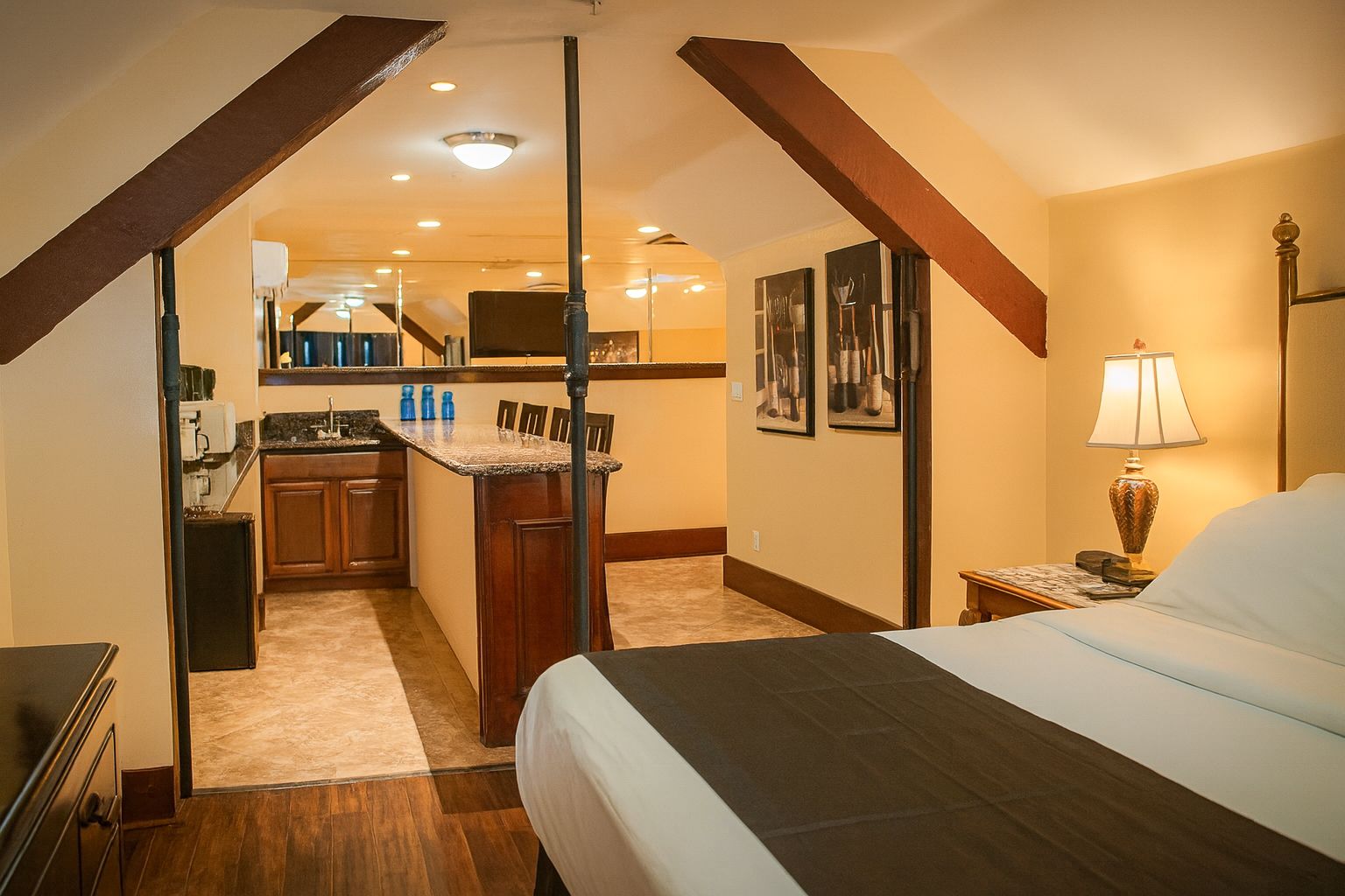 Bedroom with bed, kitchenette, and framed photos. Warm tones, wood beams, and lamp on nightstand.