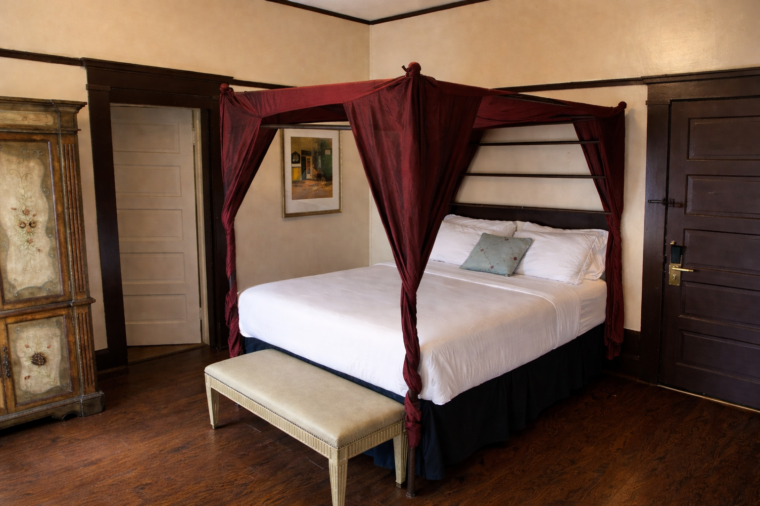 Bedroom with canopy bed, white linens, dark wood trim, bench, and antique armoire.