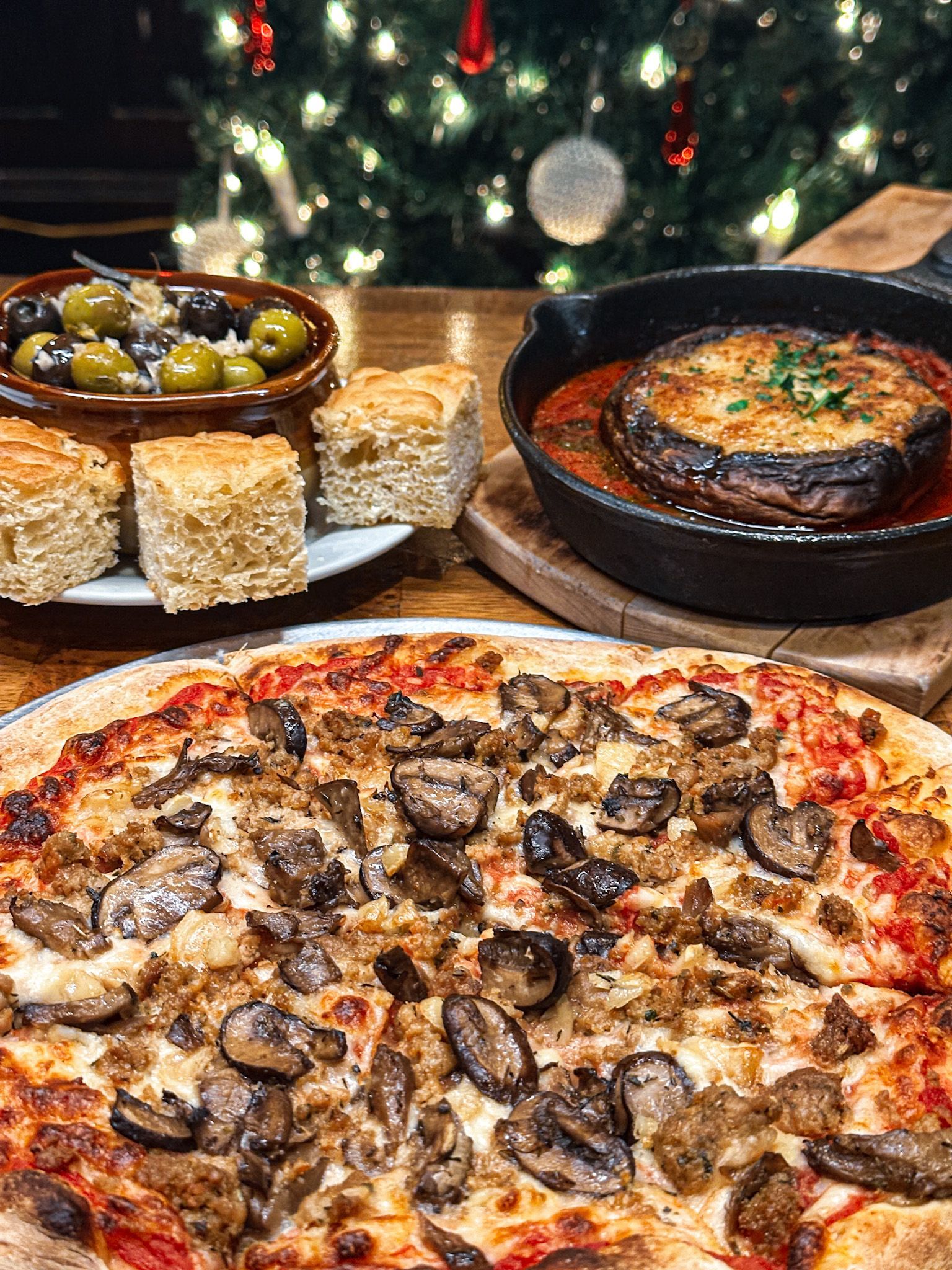 Pizza, bread, olives, and a baked mushroom dish on a table, with a Christmas tree in the background.
