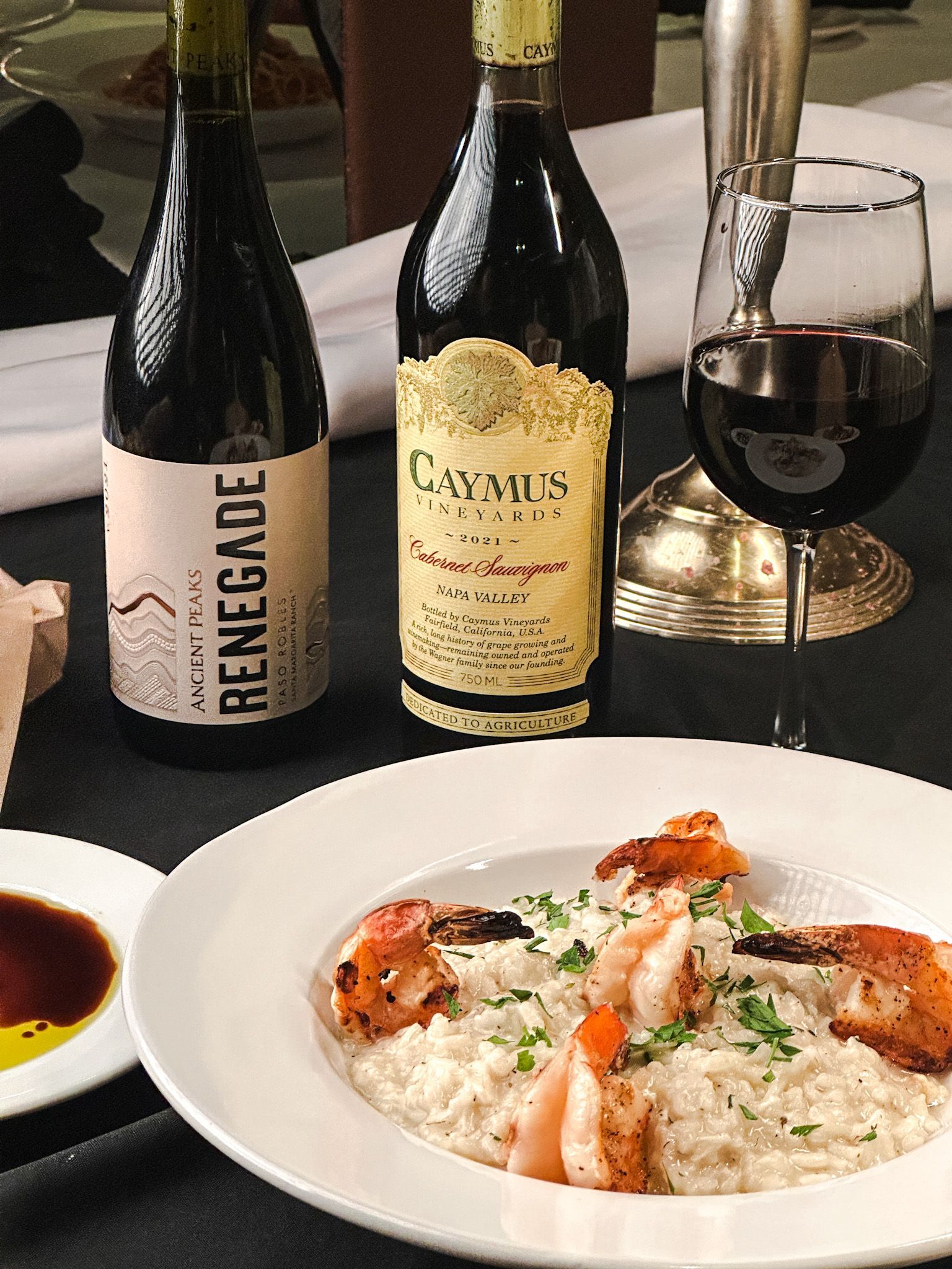 Risotto with shrimp, two wine bottles, glass of red wine, and sauce on a table.