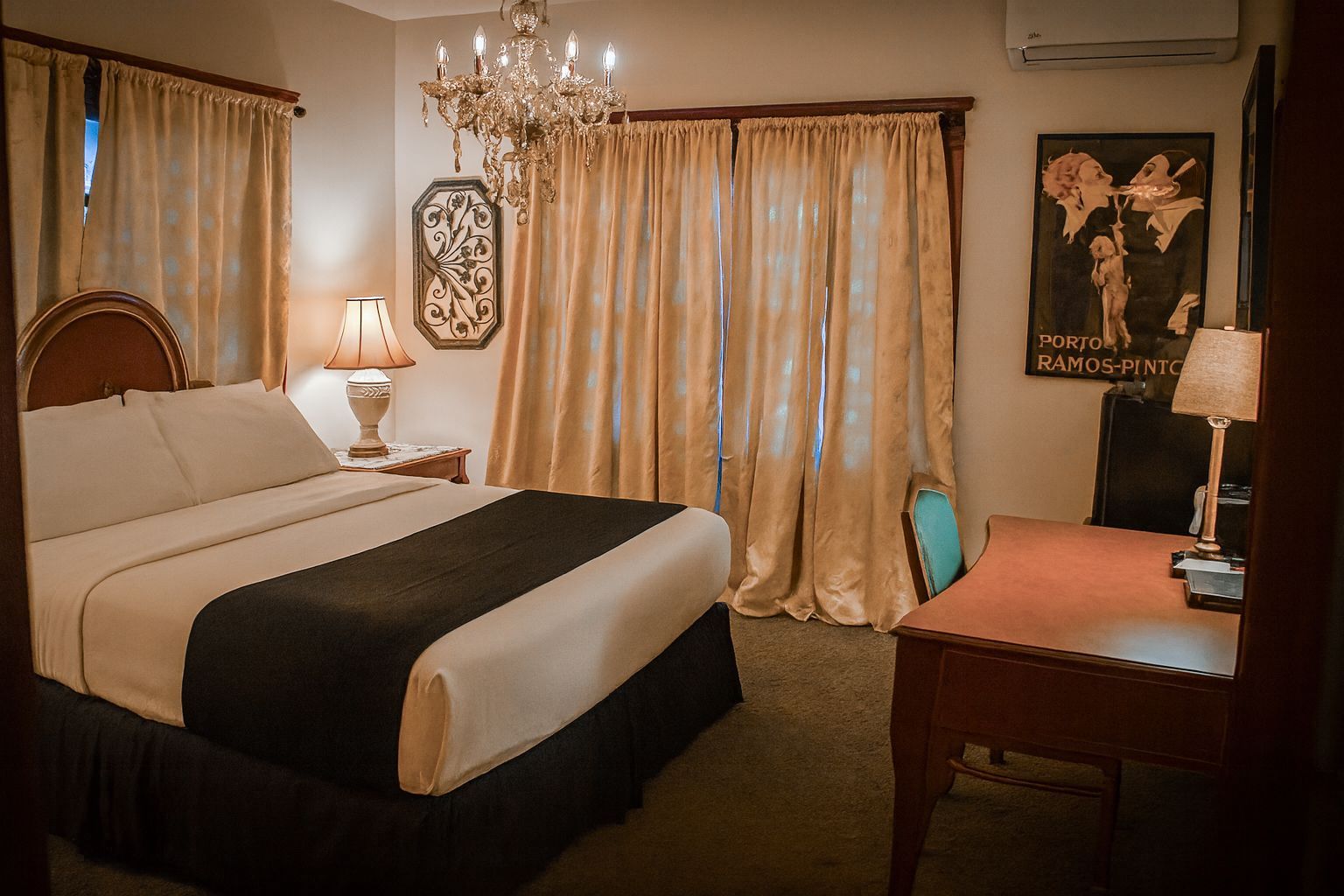 A hotel bedroom featuring a queen bed with white and dark bedding, a wooden desk, a chandelier, and gold curtains.