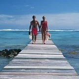Couple in red swimwear walks hand-in-hand down a wooden pier toward turquoise ocean.