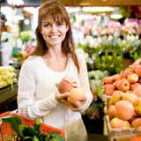 Woman in a grocery store smiles, holding two peaches. Fruit and produce surround her.