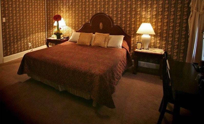 Cozy bedroom with a large bed, nightstands, and a desk. Brown patterned wallpaper and lamps illuminate the space.