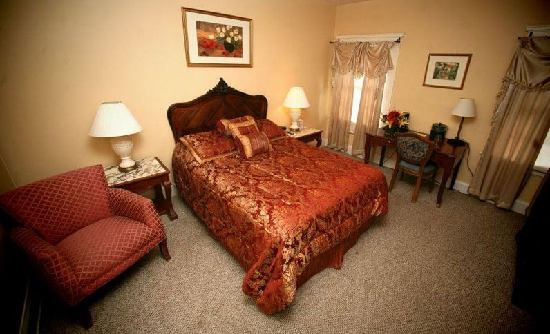 Bedroom with ornate bed, armchair, desk, and lamps. Beige walls and carpet.