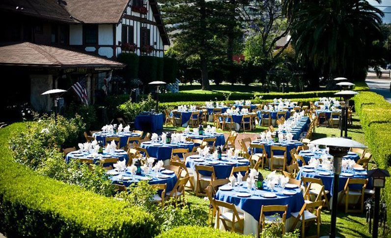 Outdoor event: Tables with blue tablecloths, set for dining, surrounded by greenery, with a Tudor-style building in the background.