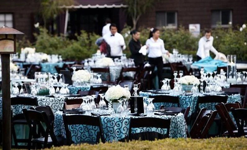 Outdoor tables set for an event with teal and black tablecloths. Servers are preparing for the event.