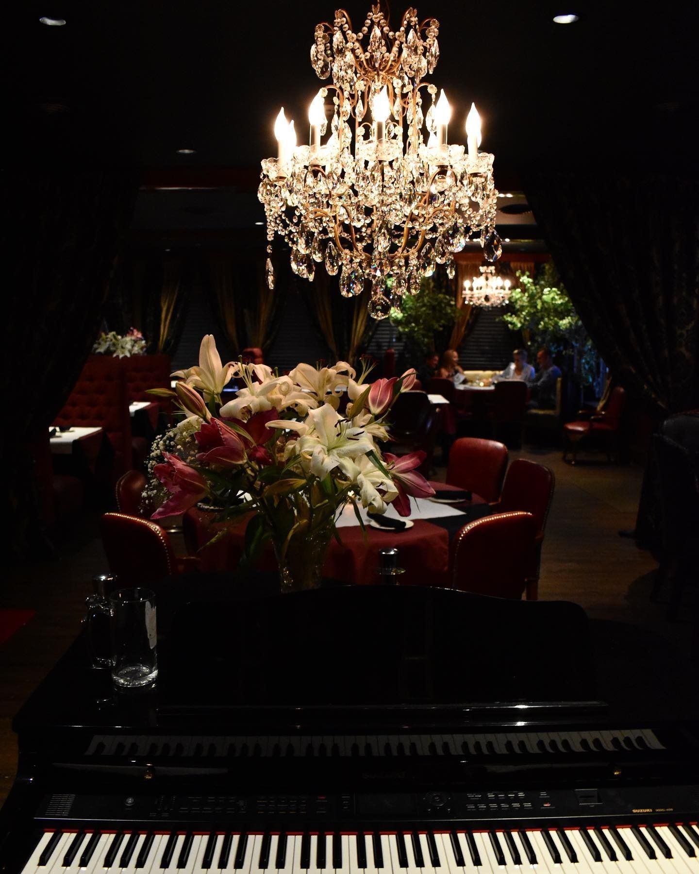 Elegant dining room with chandelier, piano, flower arrangement, and red seating.