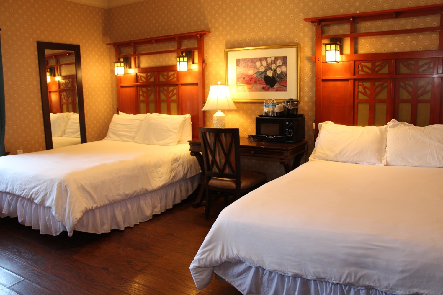 Hotel room with two beds, a desk, a mirror, and decorative headboards; warm lighting and wood flooring.