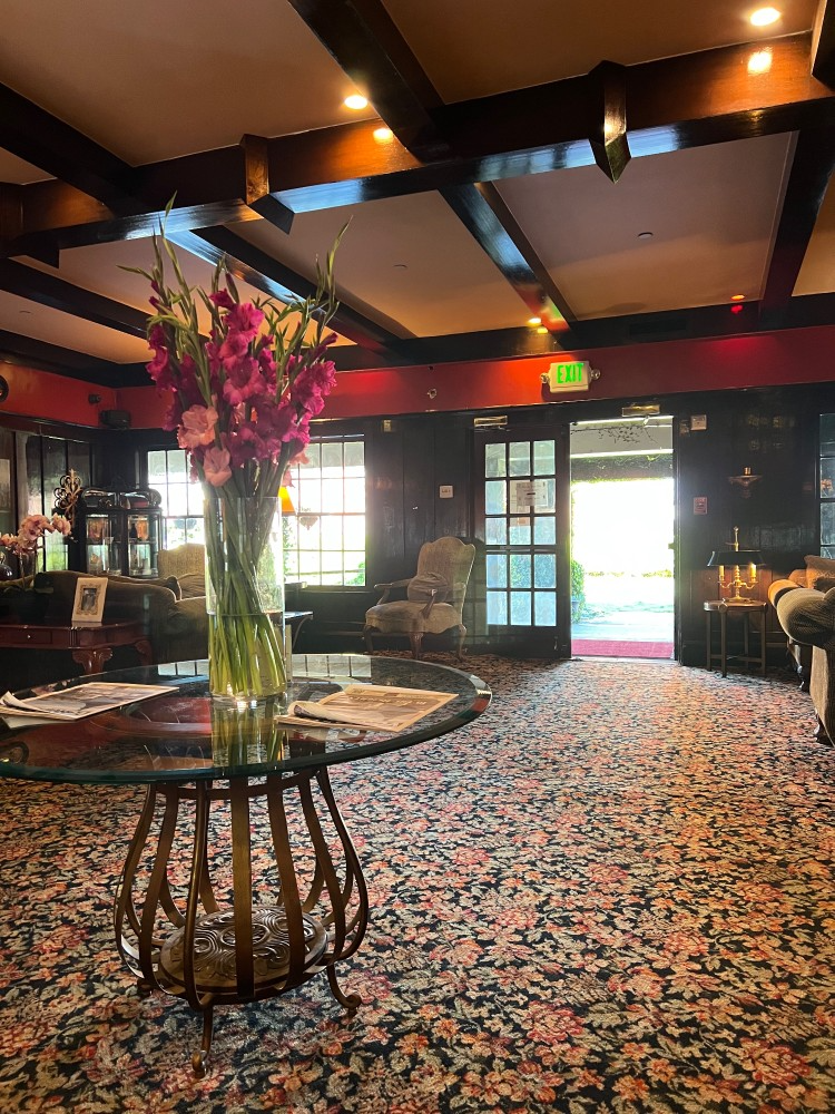 Lobby with large flower arrangement, round table, patterned carpet, wooden beams, and an open doorway.