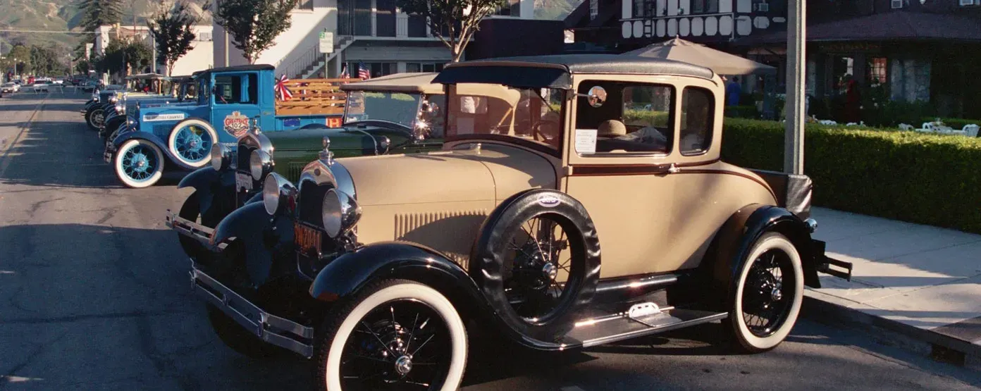 Antique cars parked on a tree-lined street with a beige Model A in the foreground.