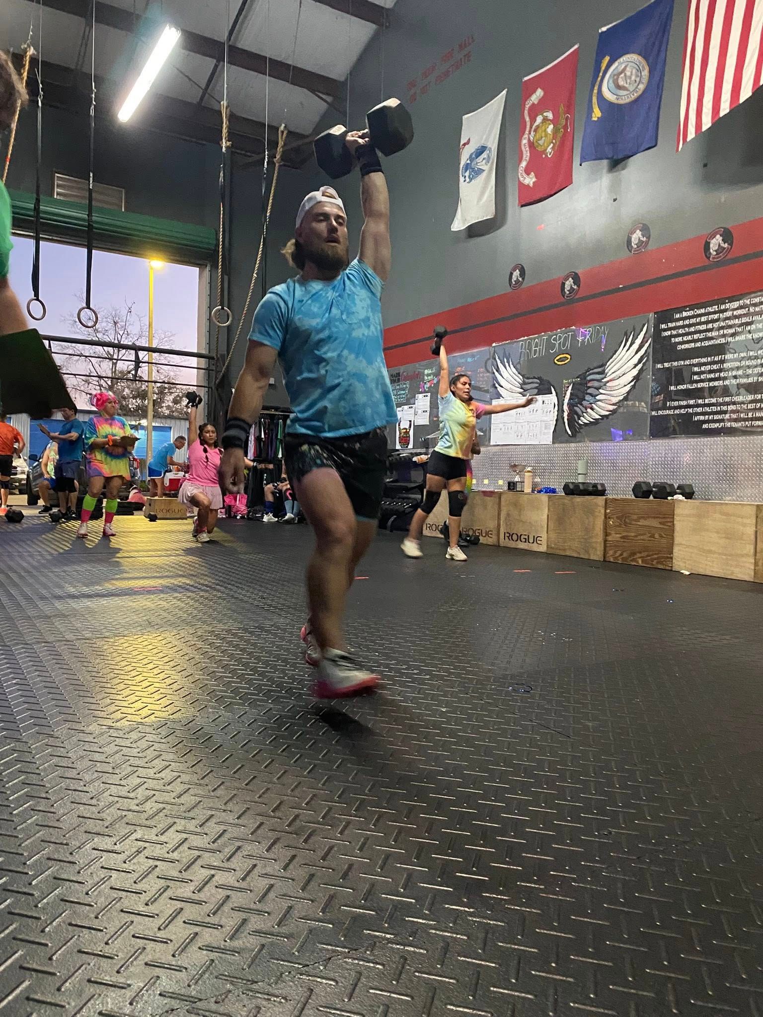 A person in a blue shirt performs a dumbbell overhead press in a gym with American and military flags on the wall.