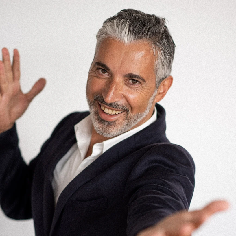 Homme souriant aux cheveux grisonnants, chemise blanche ouverte, blazer bleu marine, bras tendus, fond neutre.