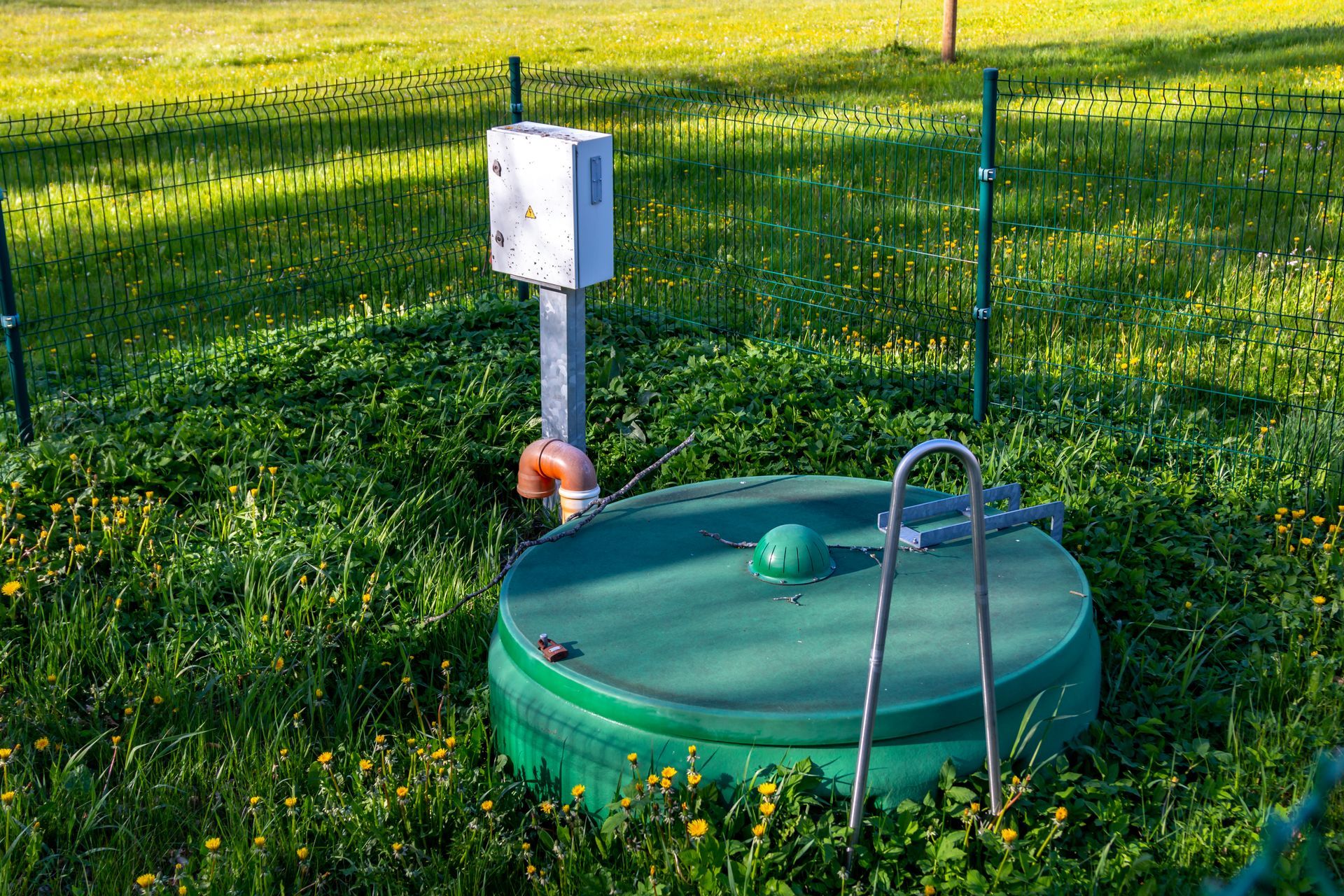 Green septic tank with metal handle in grassy yard, with electrical box and pipes.