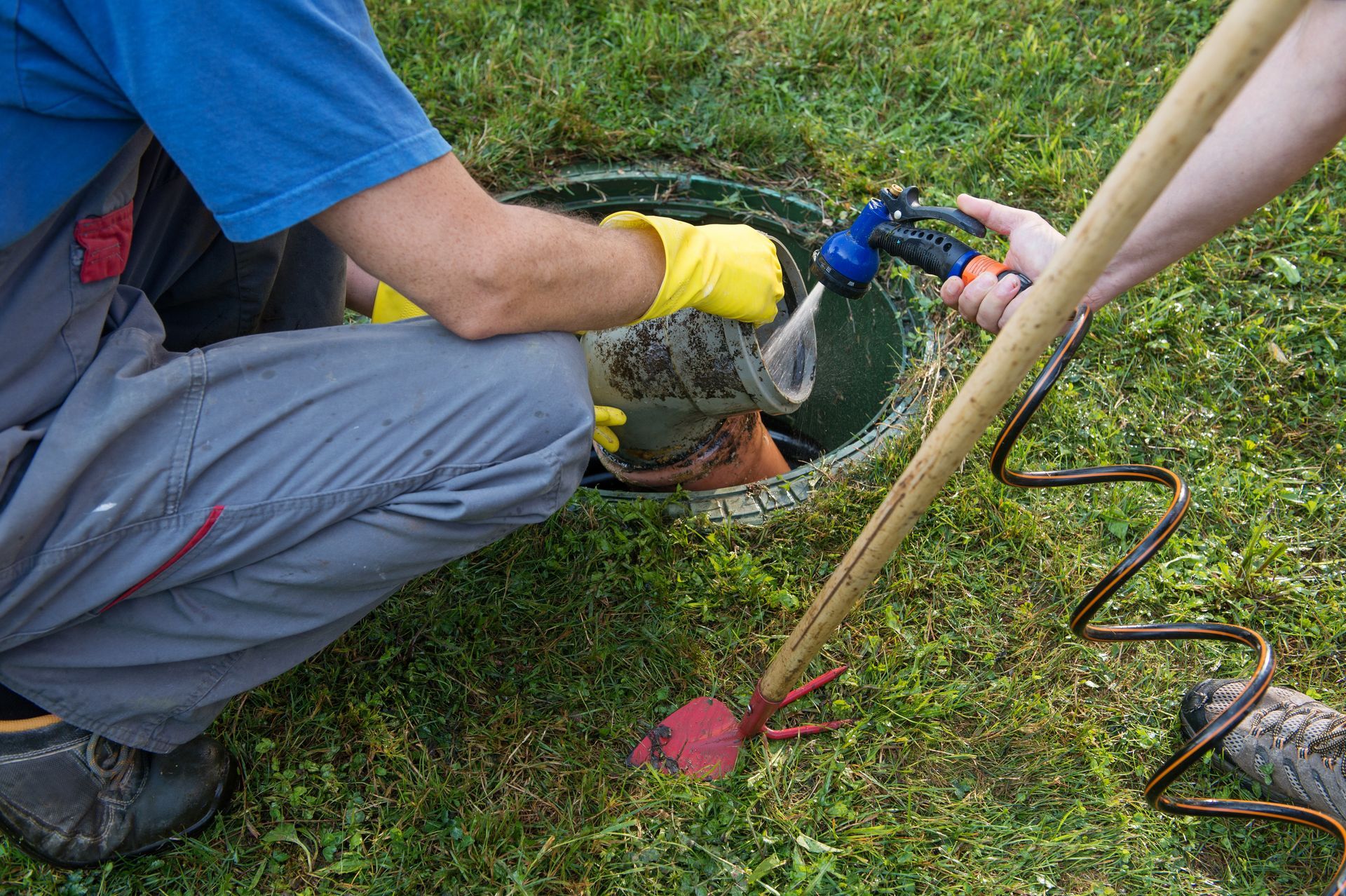 A man wearing yellow gloves is kneeling in the grass next to a manhole cover.