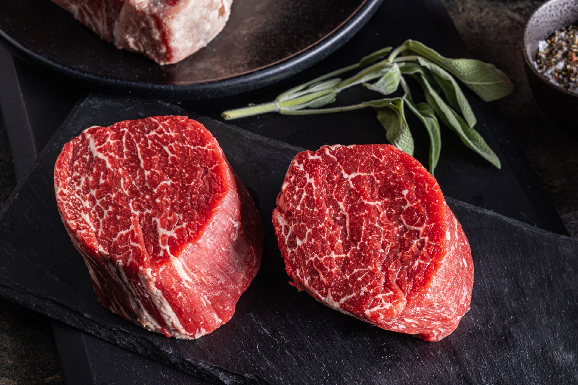 Two steaks are sitting on a black cutting board next to a plate of meat.