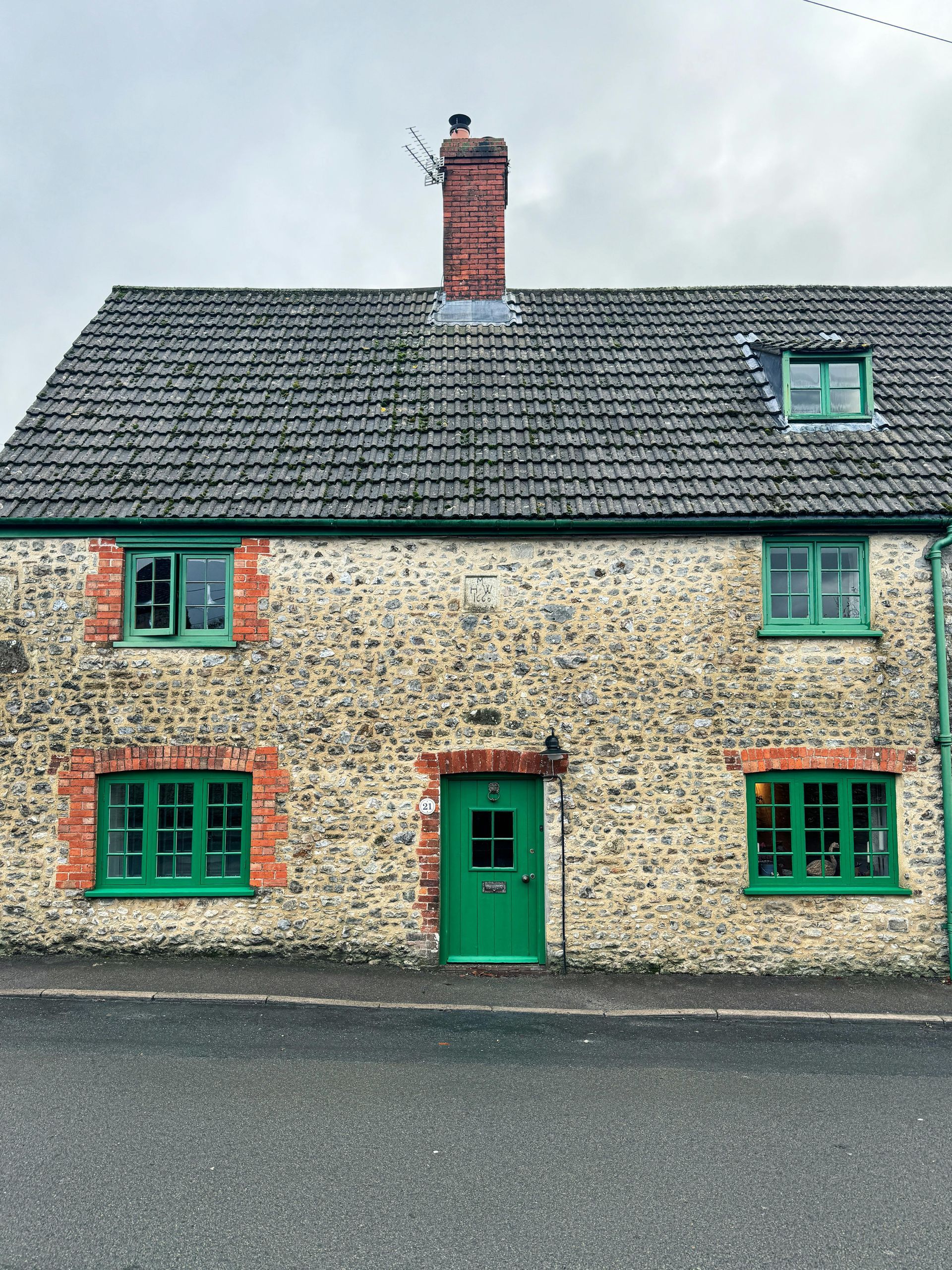 Stone cottage with green door and window frames; brick accents. Gray tile roof; overcast sky.