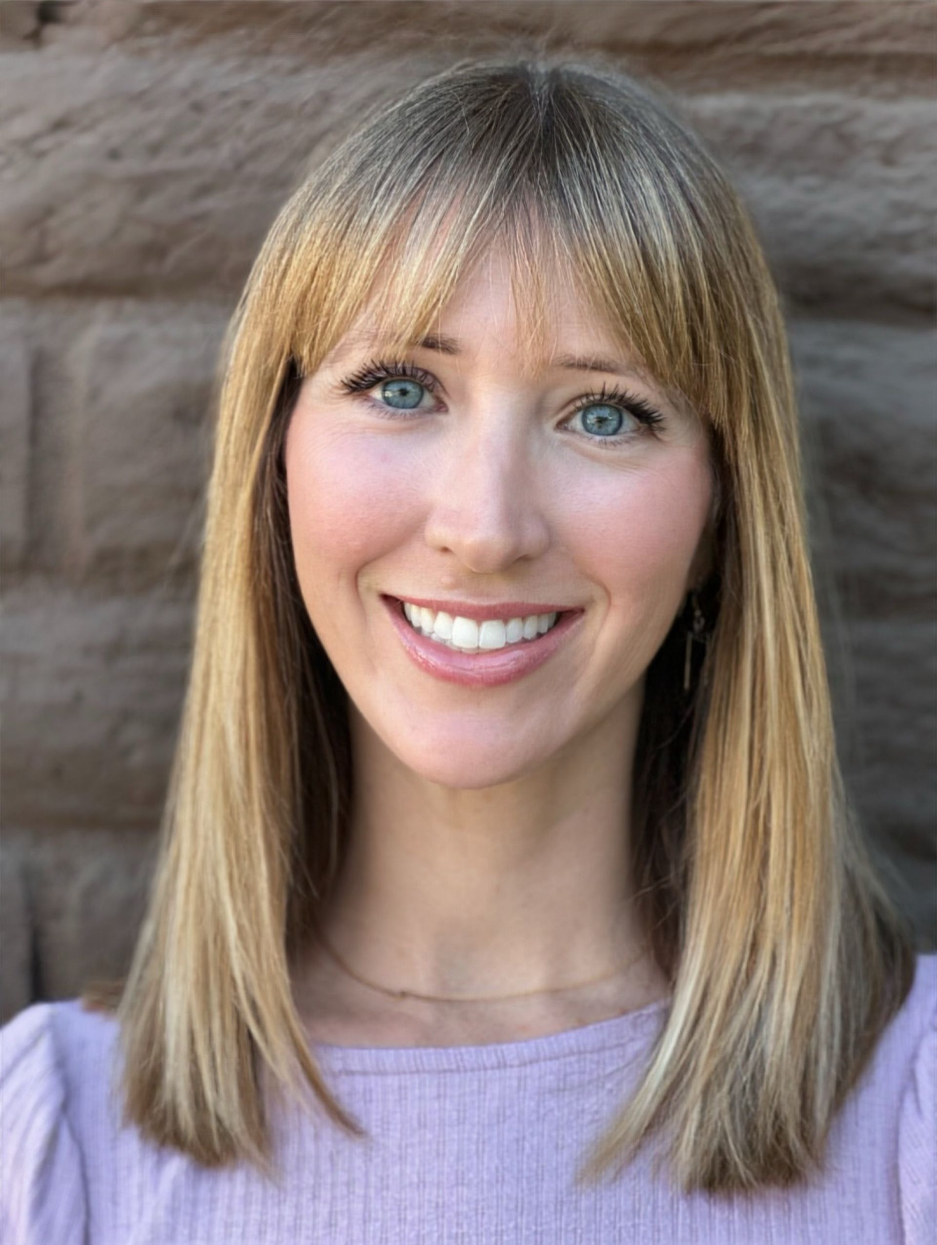 Woman with blonde hair and bangs smiles, in front of a brick wall.