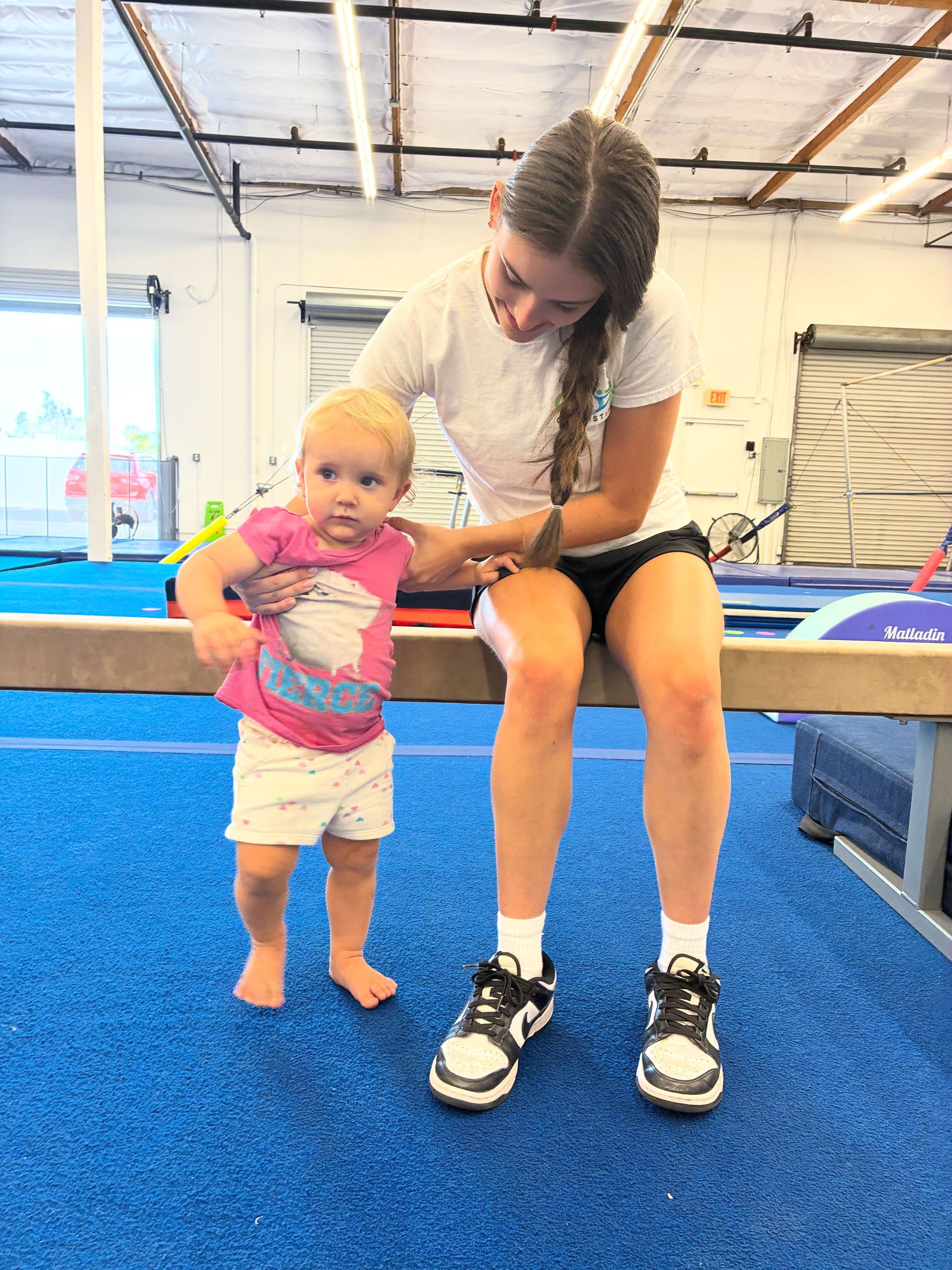 Baby learning to walk with a woman in a gymnastics studio, blue floor.