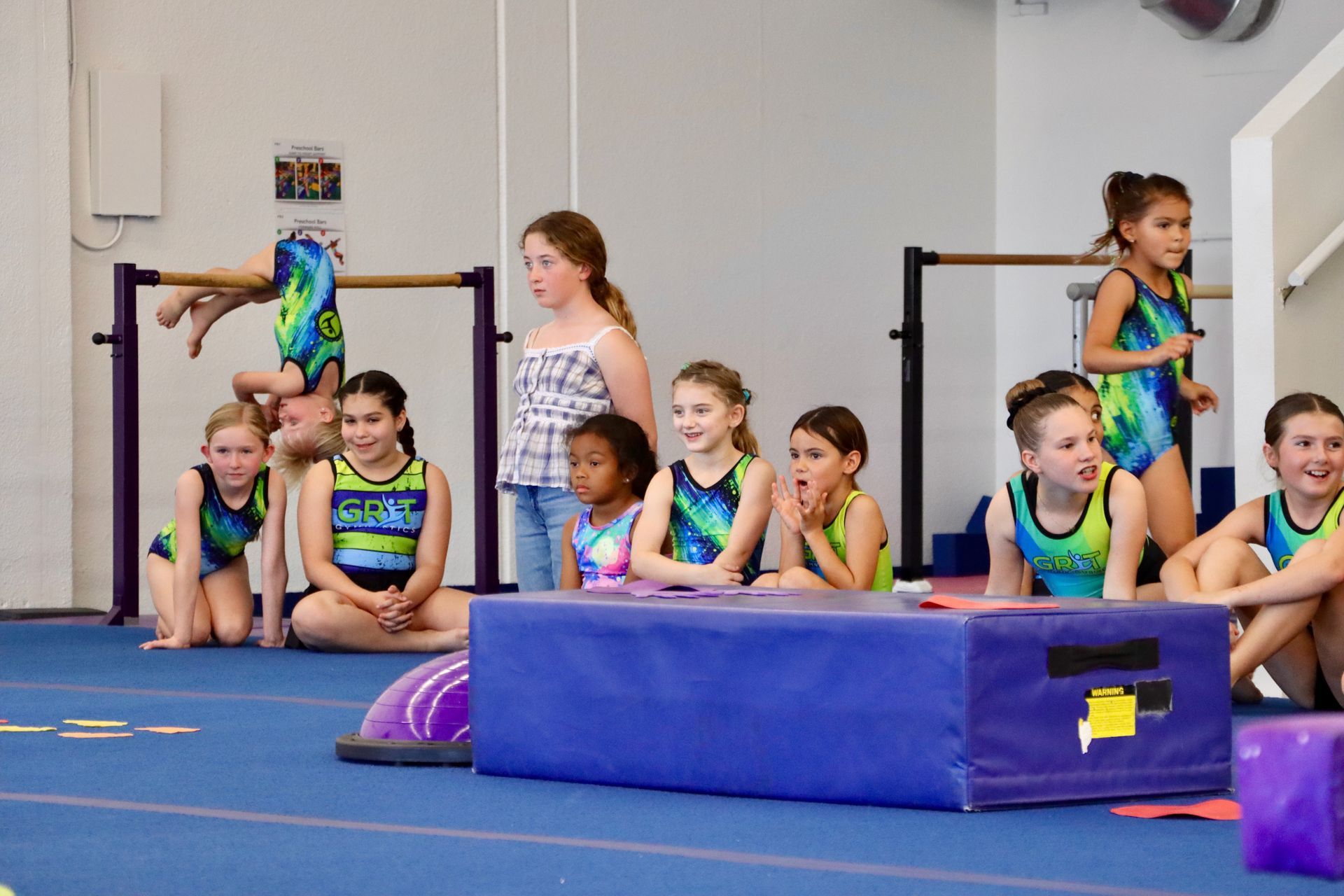 Gymnastics class: Kids watch a girl on bars. Others in blue leotards sit, standing by blue mats.
