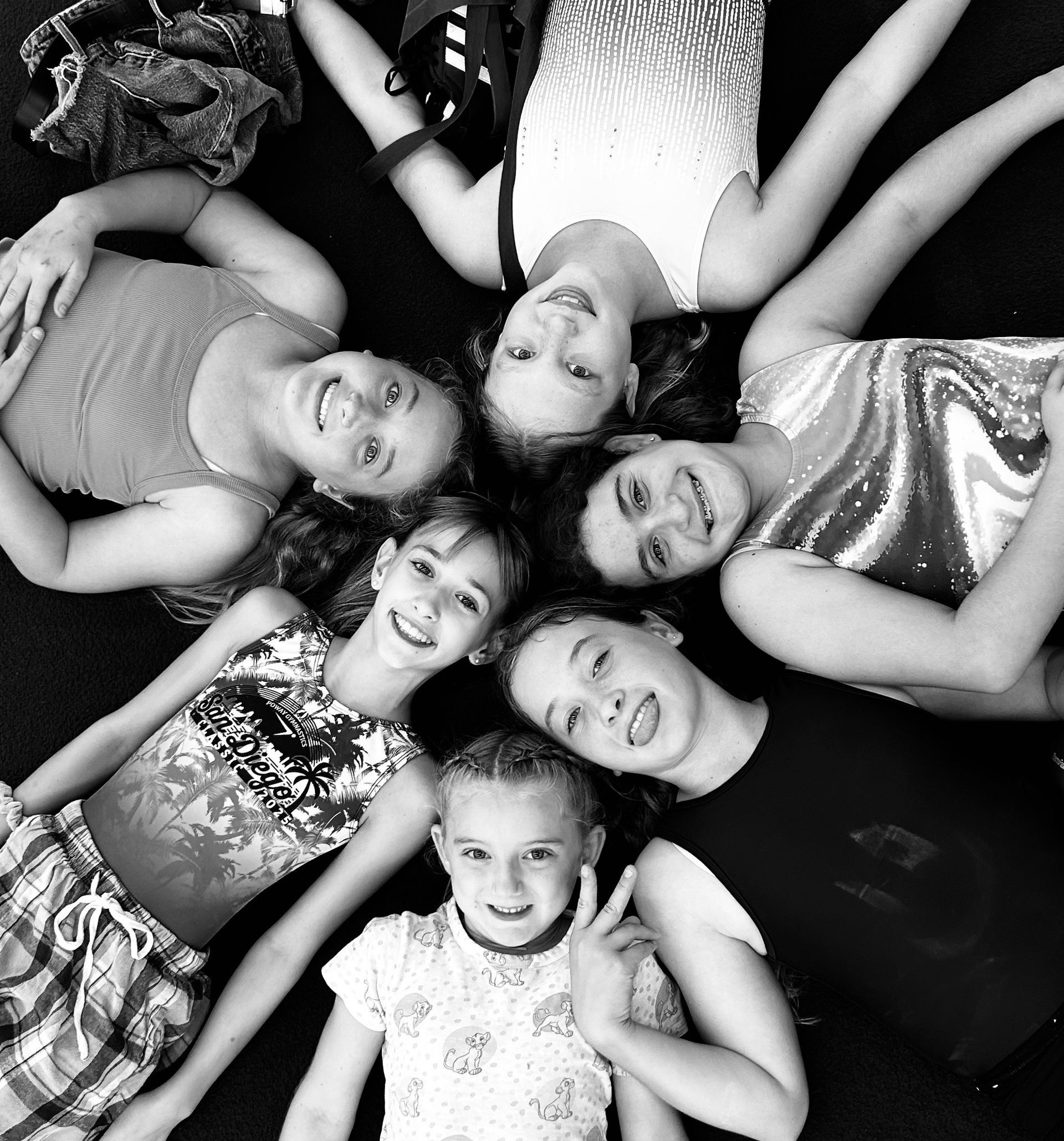 Group of smiling people lying in a circle, arms outstretched, on a dark surface.