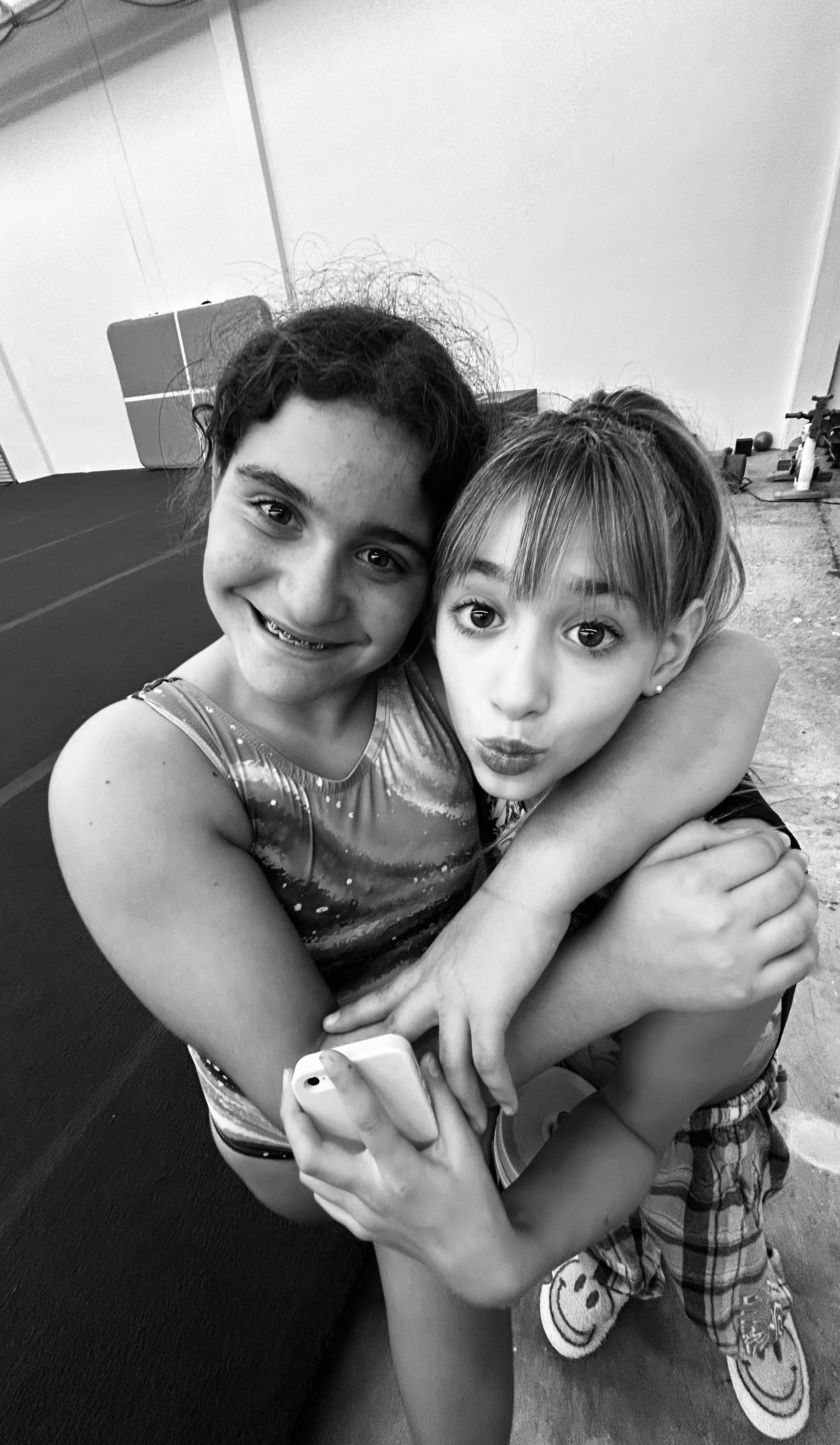 Two girls hugging, smiling. One holds a phone. Gymnastics attire, inside a gym.