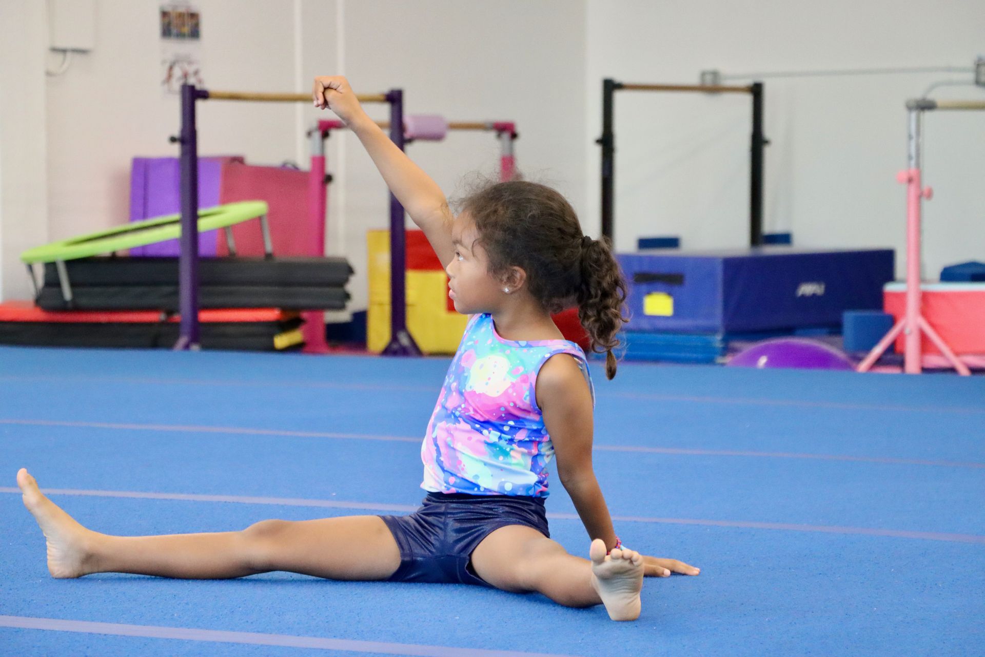 Girl on balance beam, another girl spots her. Gym setting, red beam, blue mat.