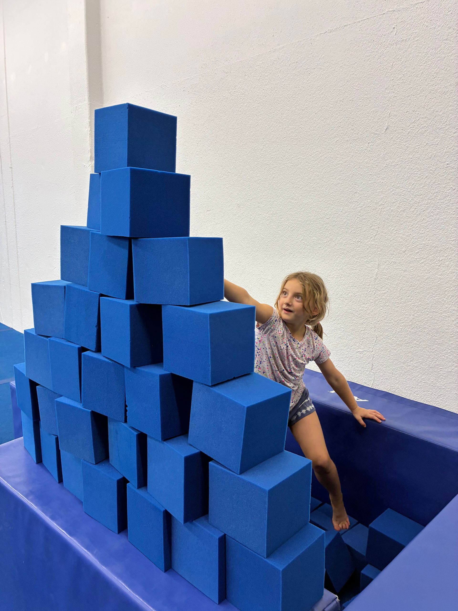 A child climbs a stack of blue foam blocks inside a play area.