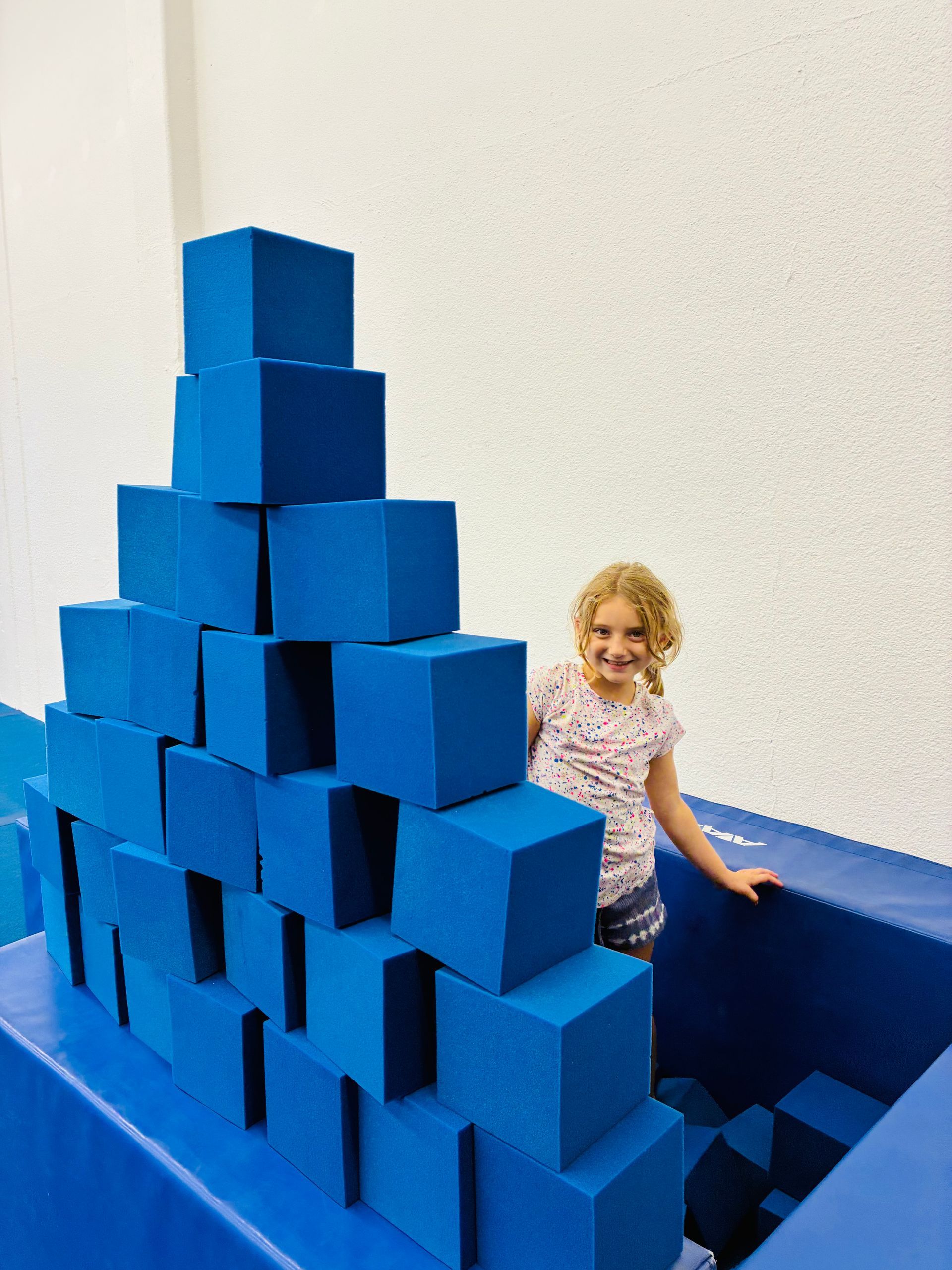 Girl smiling next to a tall blue foam block structure in a play area.