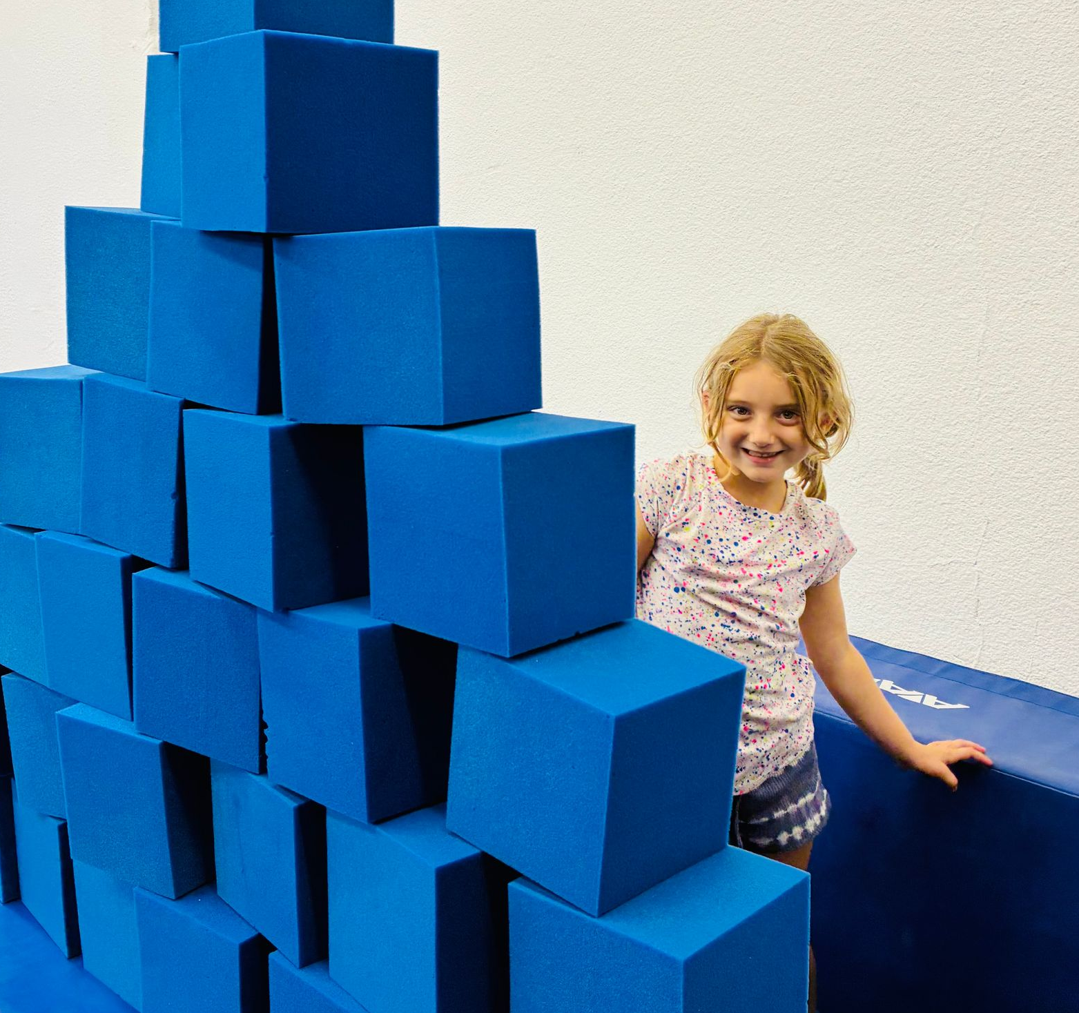 Girl smiles next to a blue cube structure in a play area.