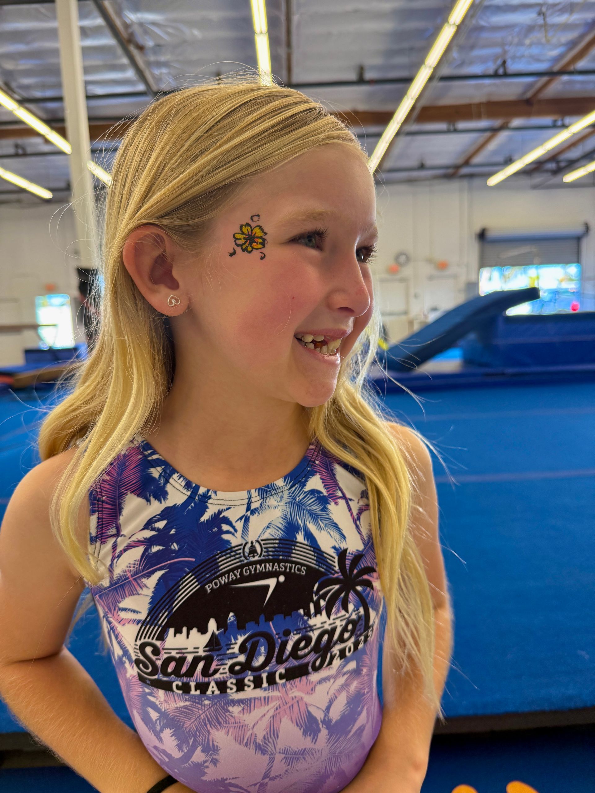 Young girl with blonde hair, a bee face painting, and a San Diego leotard, smiles in a gym.