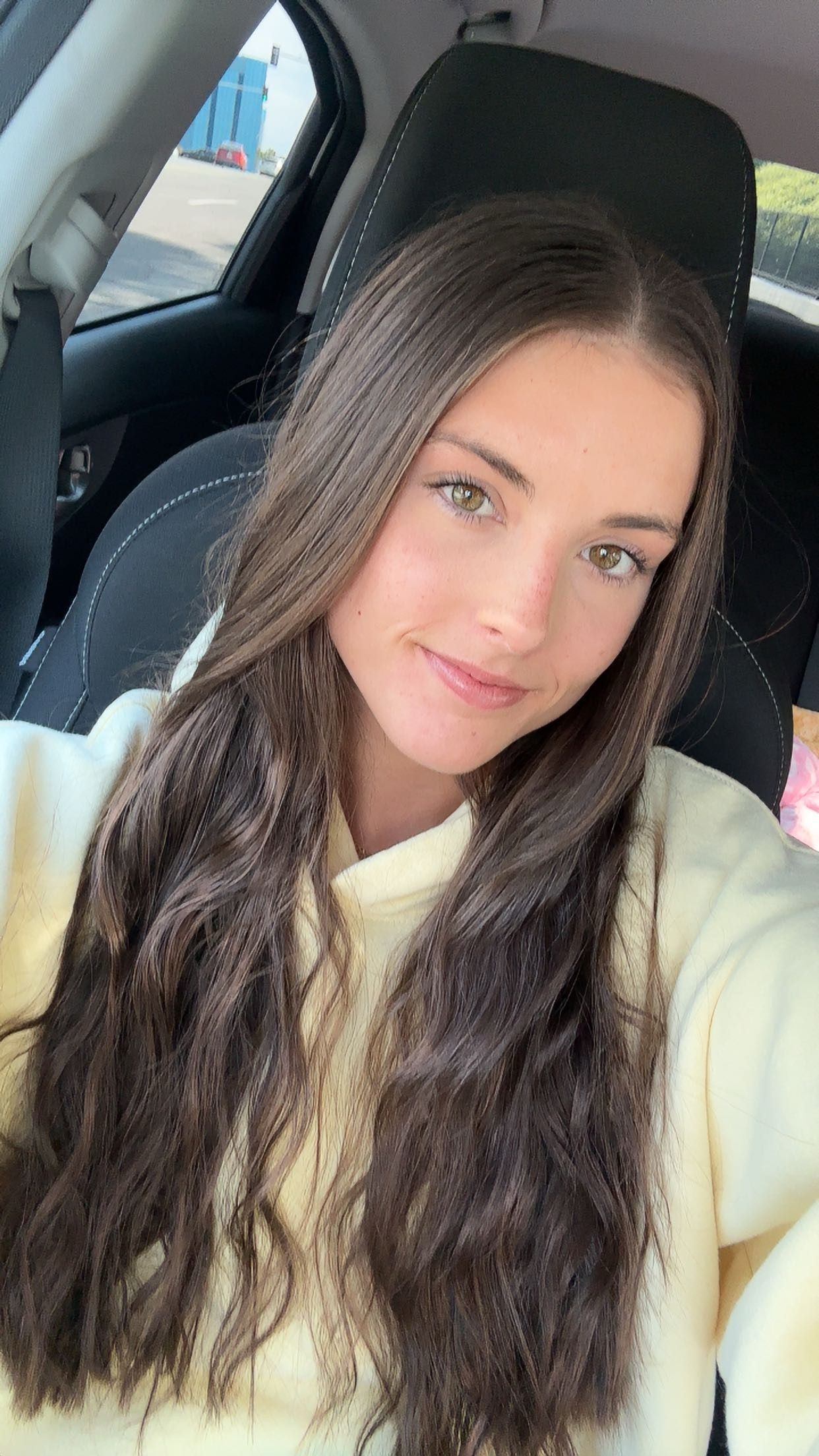 Woman with long brown hair in a car, smiling slightly. Wearing a light yellow sweater.