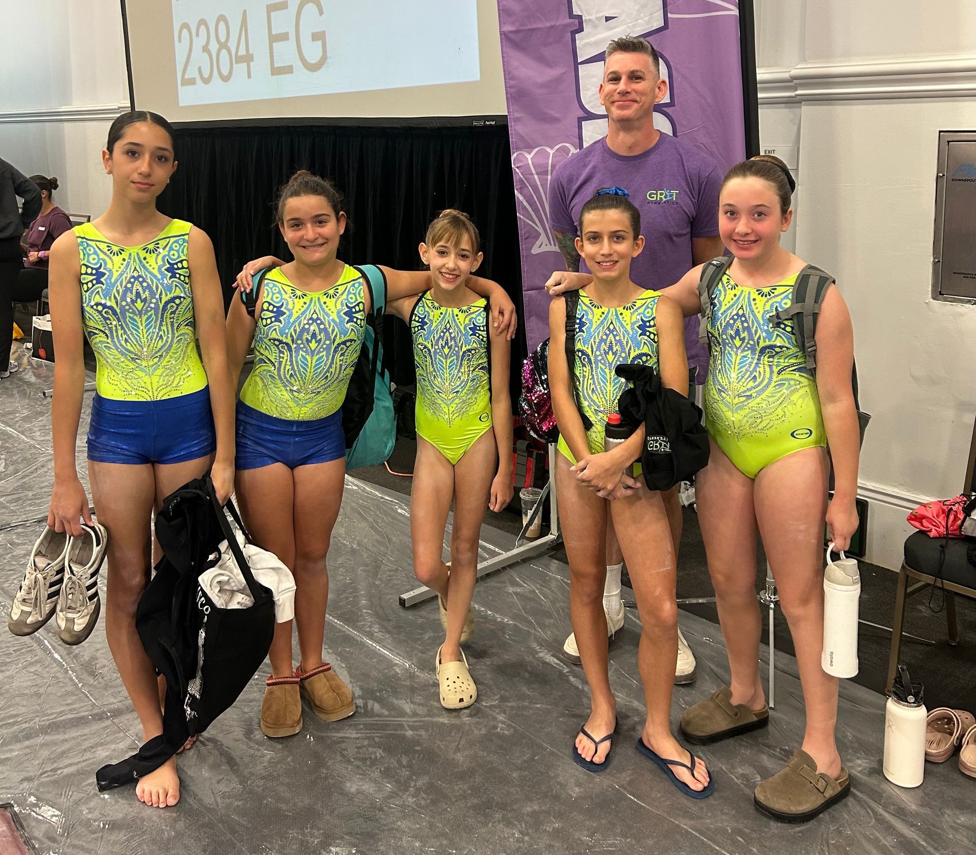 Gymnast performing a vault, arms outstretched, mid-air. Red vault, blue and white leotard. Spectators in background.