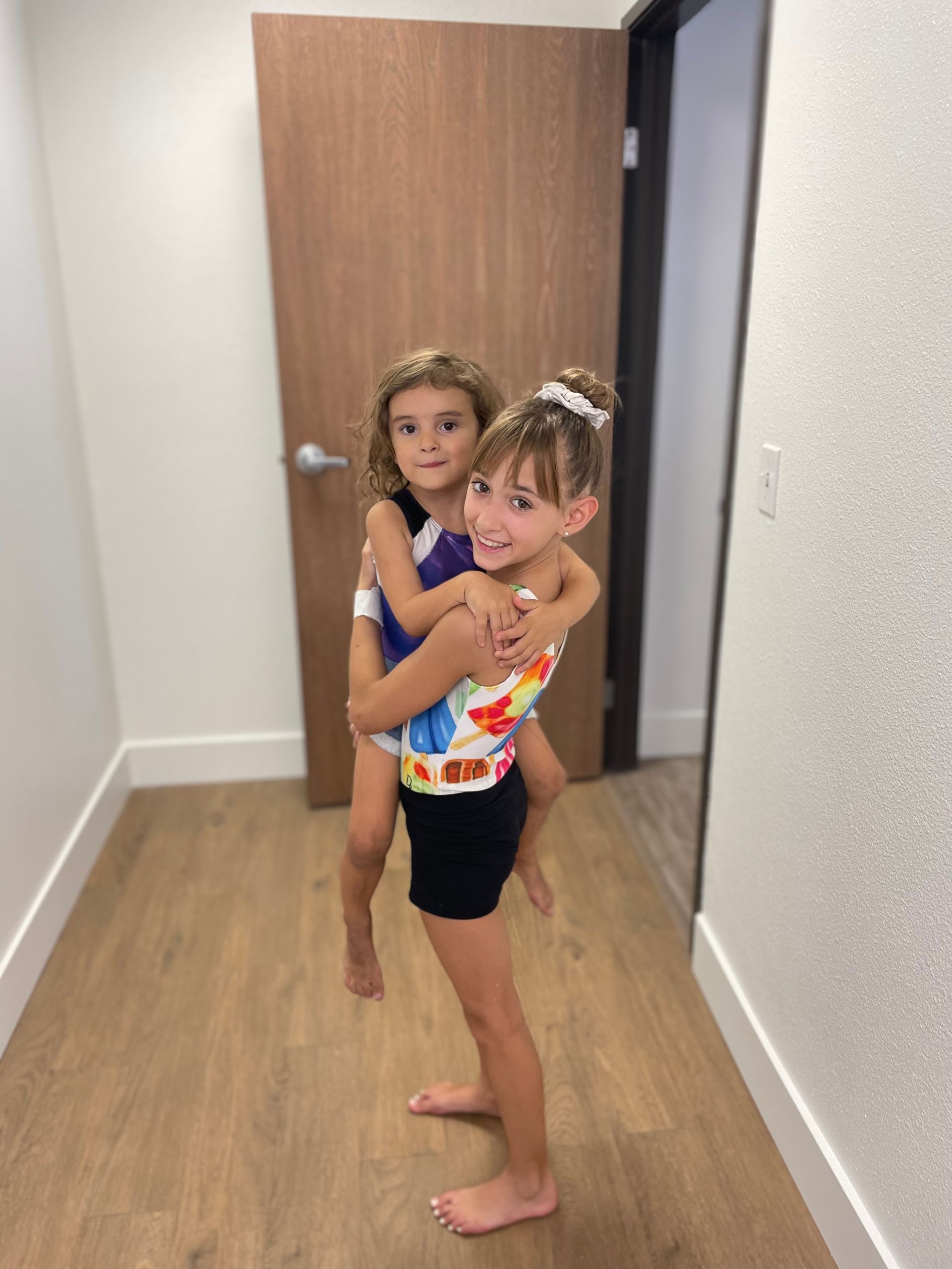 Girl on balance beam, another girl spots her. Gym setting, red beam, blue mat.