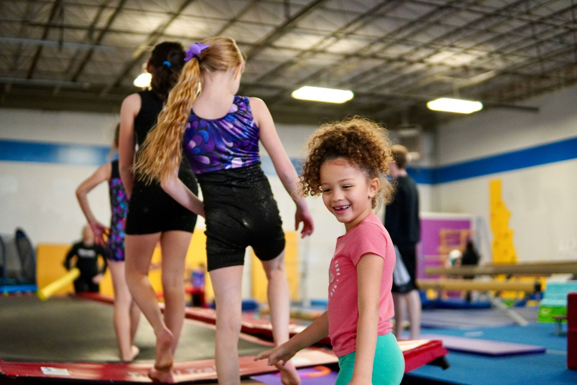 Young girl smiles at the camera while walking on a balance beam in a gymnastics gym. Other girls in leotards are nearby.