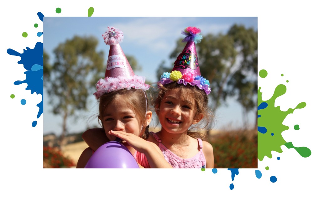 Two girls in party hats, one blowing up a purple balloon, smiling outdoors.