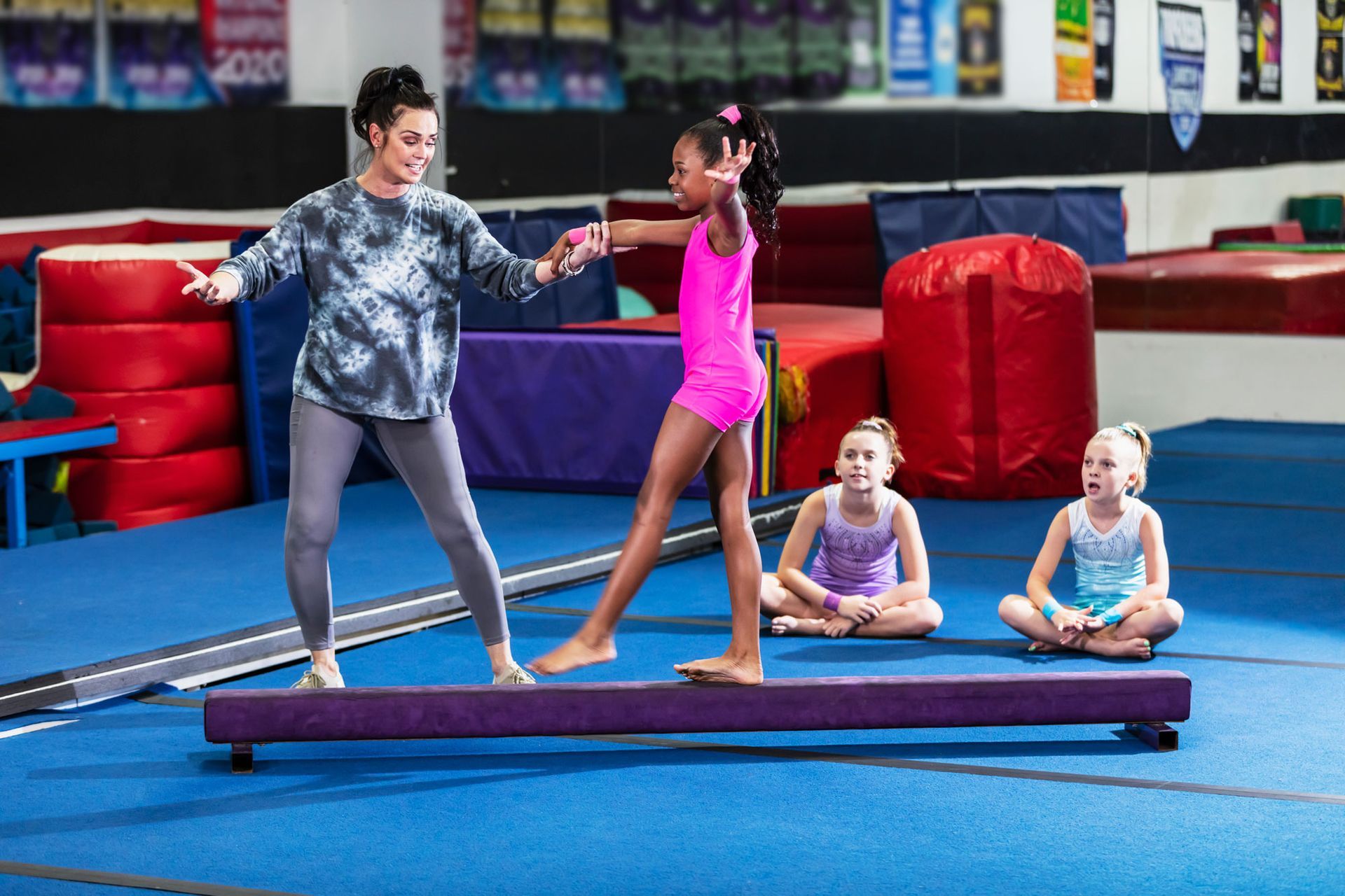 A woman helps a young girl balance on a low balance beam in a gymnastics gym, two other girls watch.