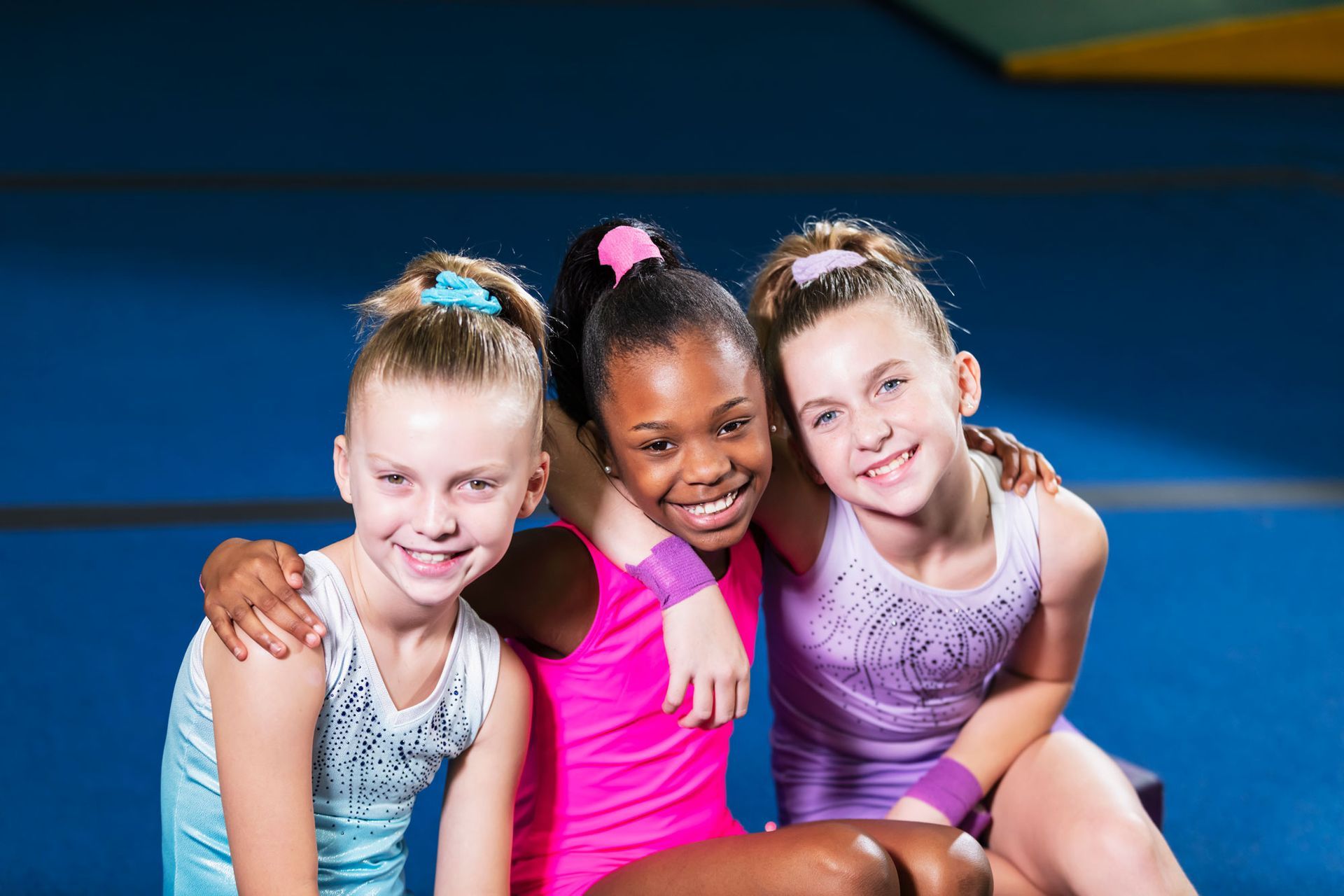 Three gymnasts in athletic wear smile, arm-in-arm. Bright pink, light blue, and lavender leotards. Blue gym setting.