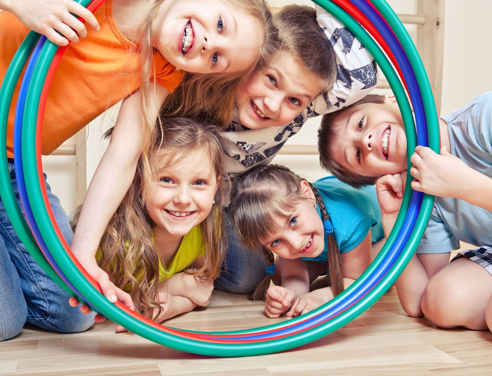 Children smiling while holding hula hoops, indoors, on a wooden floor. They are looking through the hoops toward the viewer.