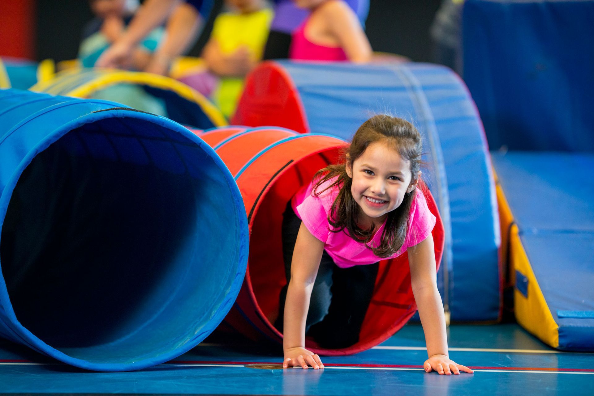 Girl crawls through a red tunnel at an indoor gym. She smiles while looking at the camera.