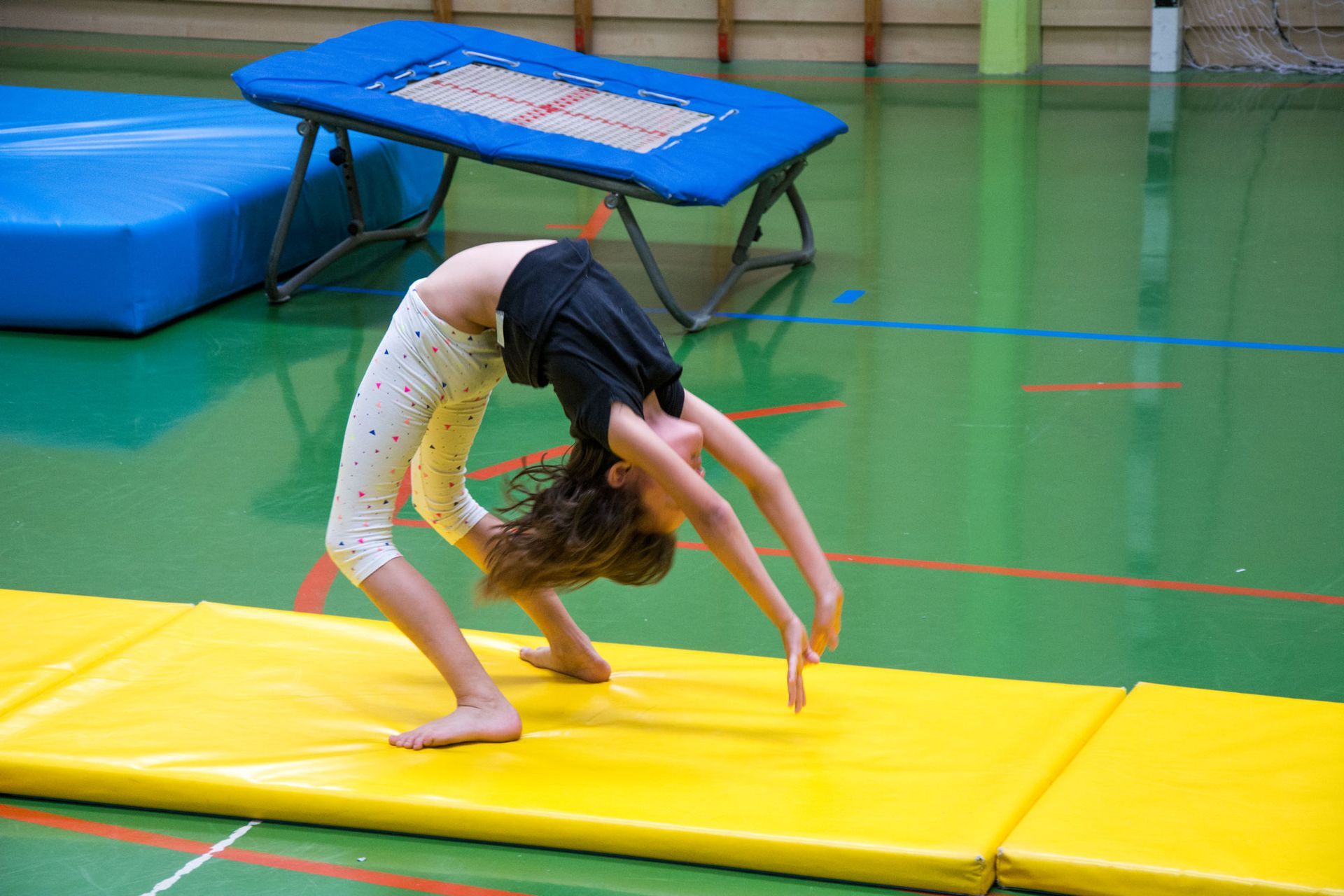 A young girl in a black shirt and polka-dot leggings performs a backbend on a yellow mat in a gym.