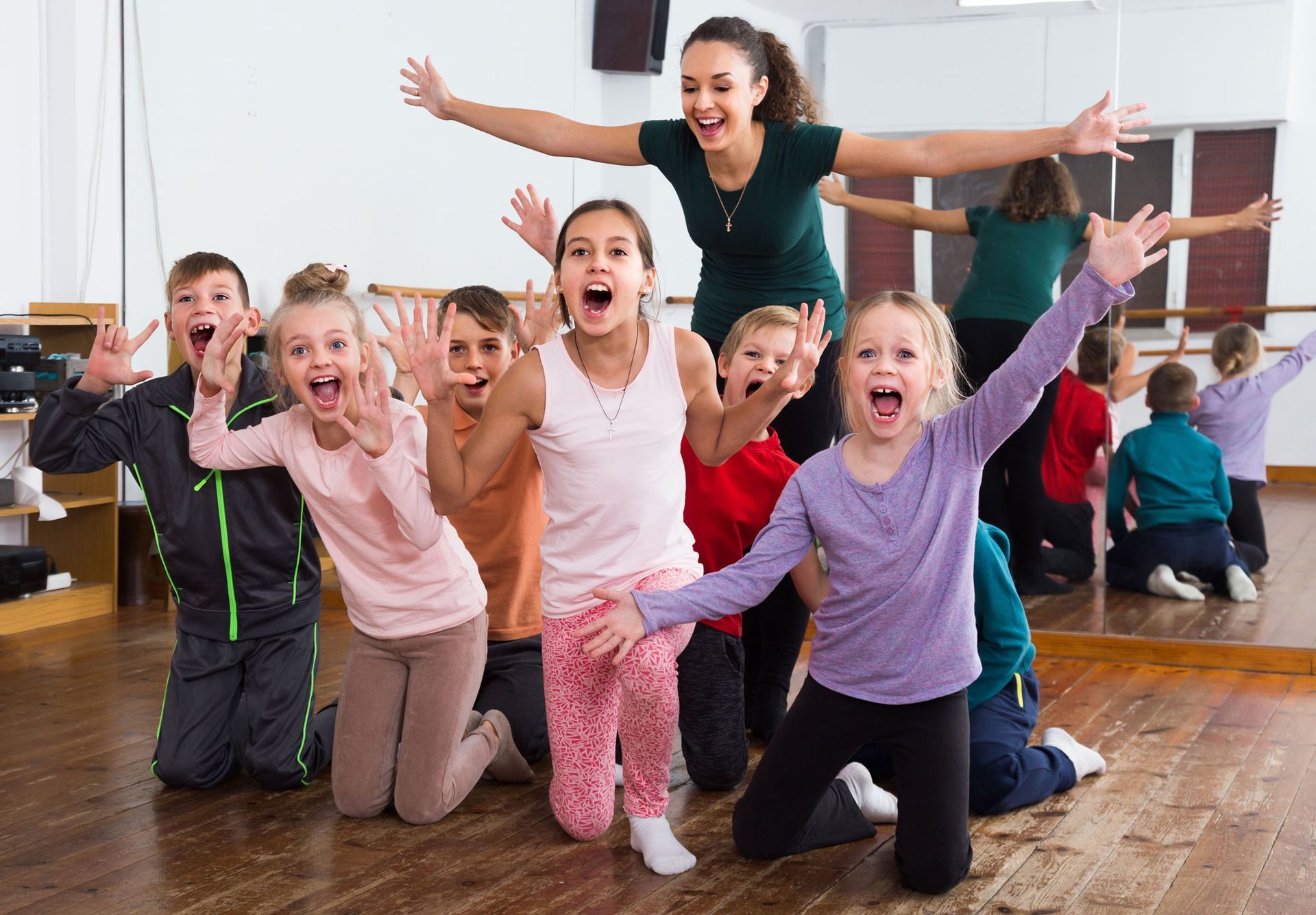 Group of children and a teacher in a dance studio, all enthusiastically posing with excited expressions and outstretched arms.