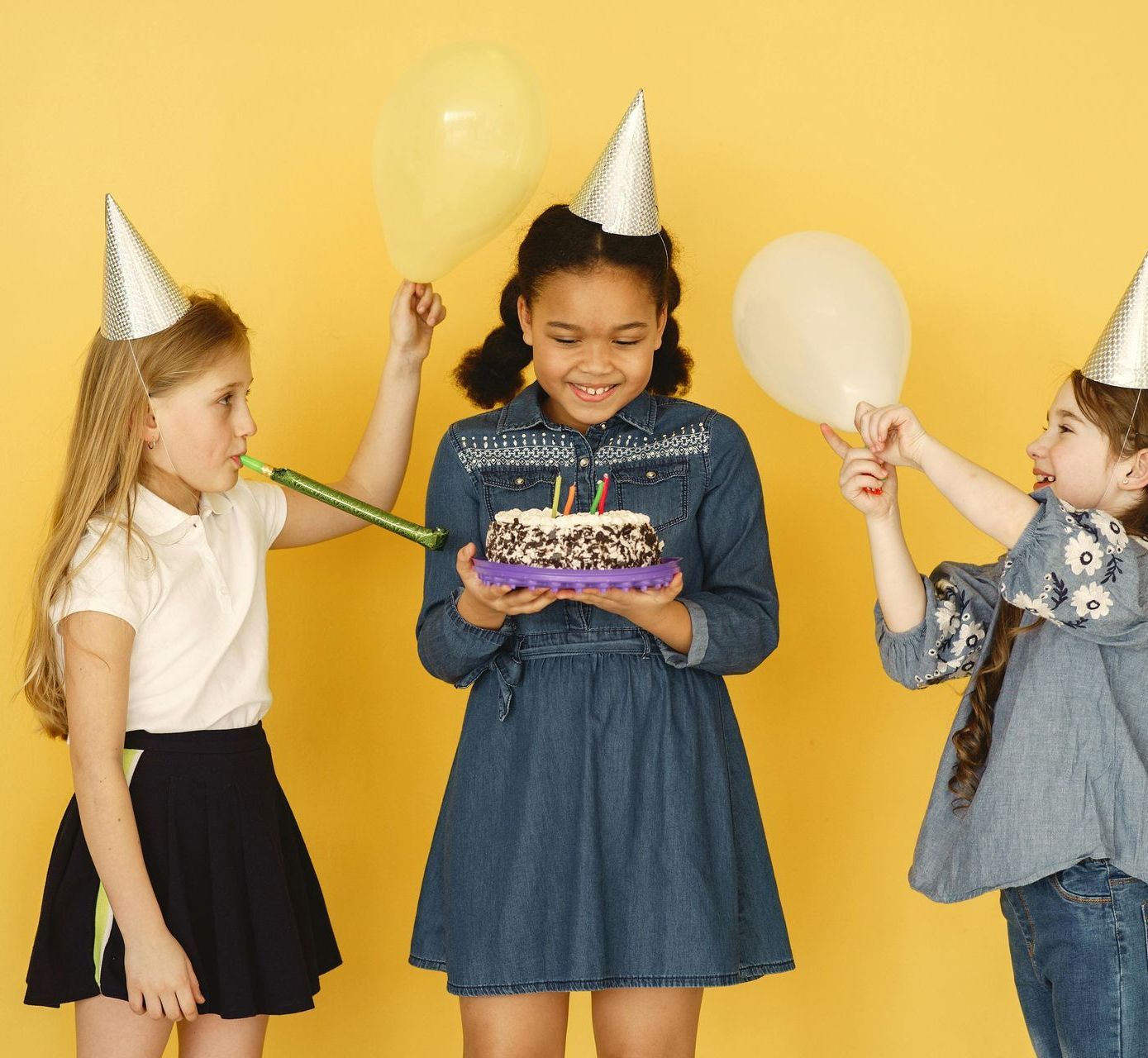 Three young girls celebrate a birthday. One holds a cake, two wear party hats, and all are smiling against a yellow backdrop.