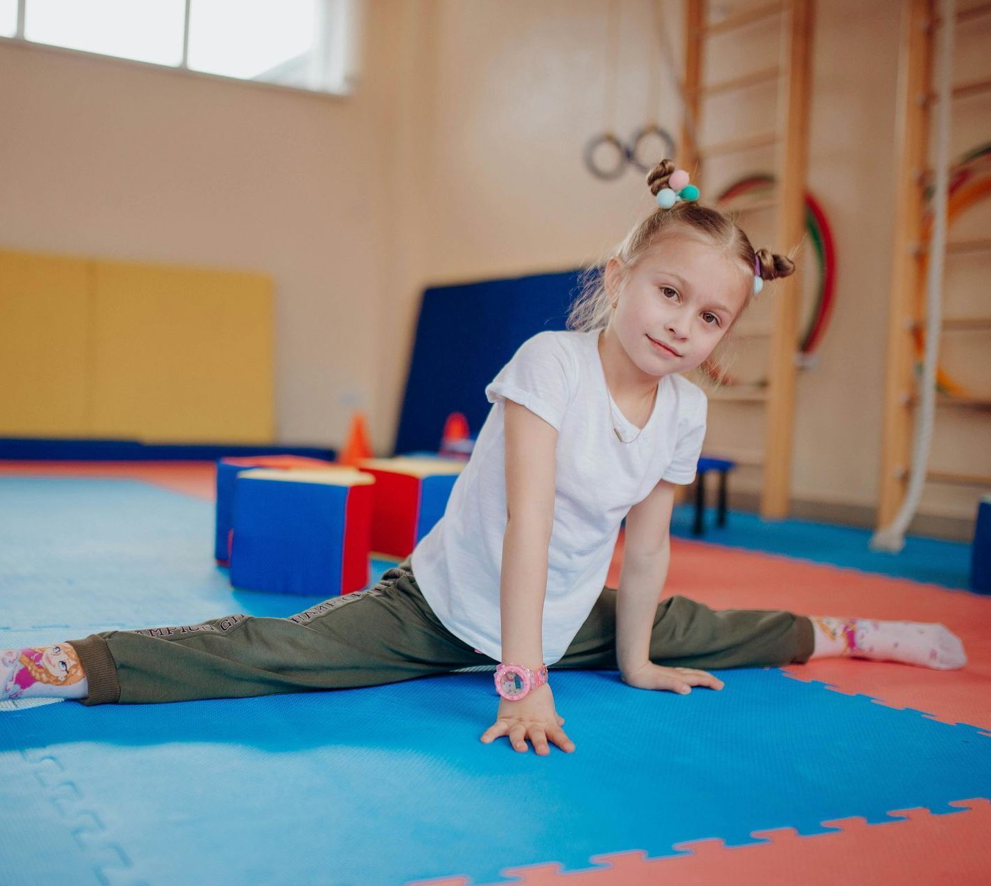 Young girl with blonde hair in pigtails, wearing a white shirt and green pants, smiles while doing the splits on a gym floor.