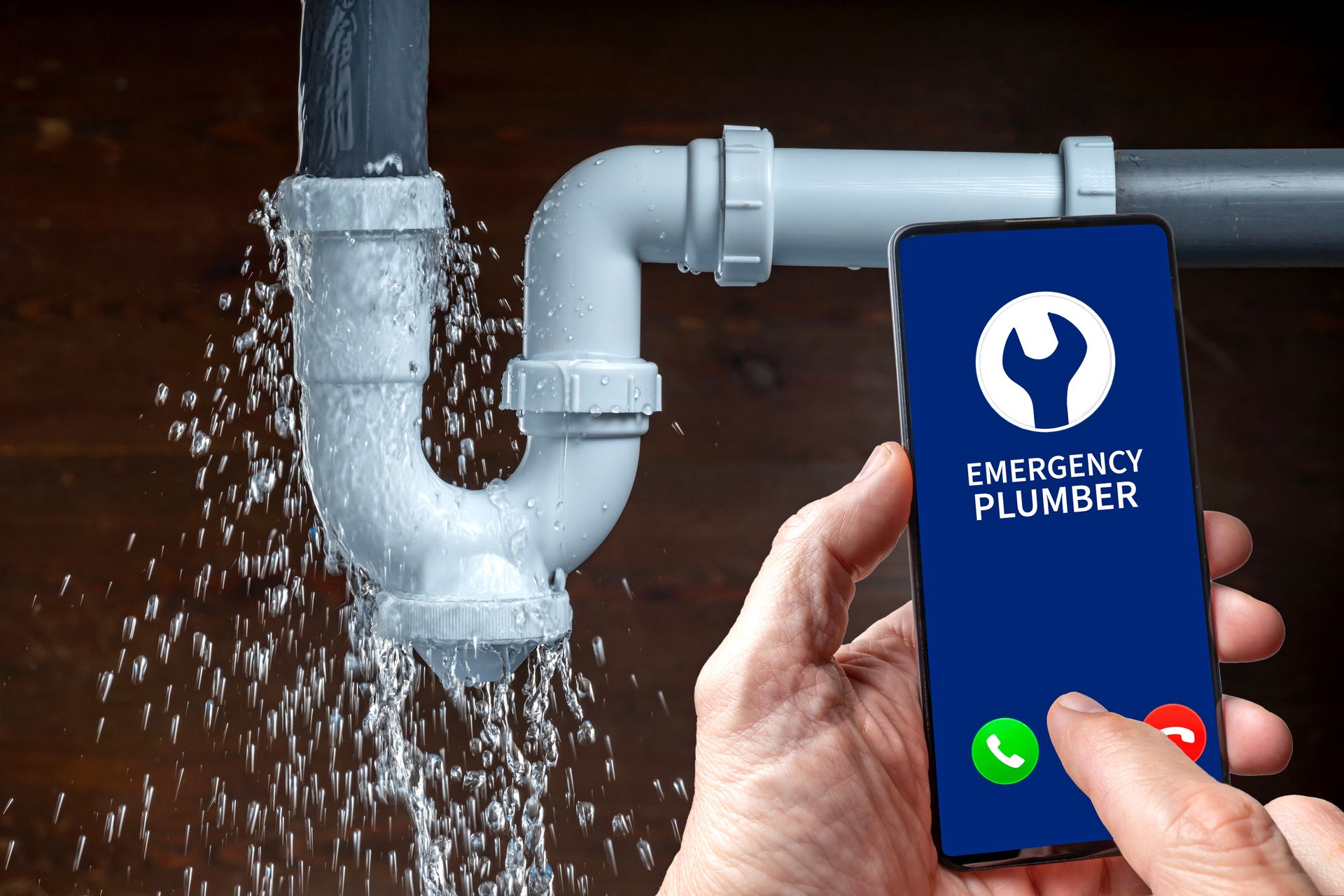 Leaking sink pipe and hand calling an emergency plumber on smartphone.